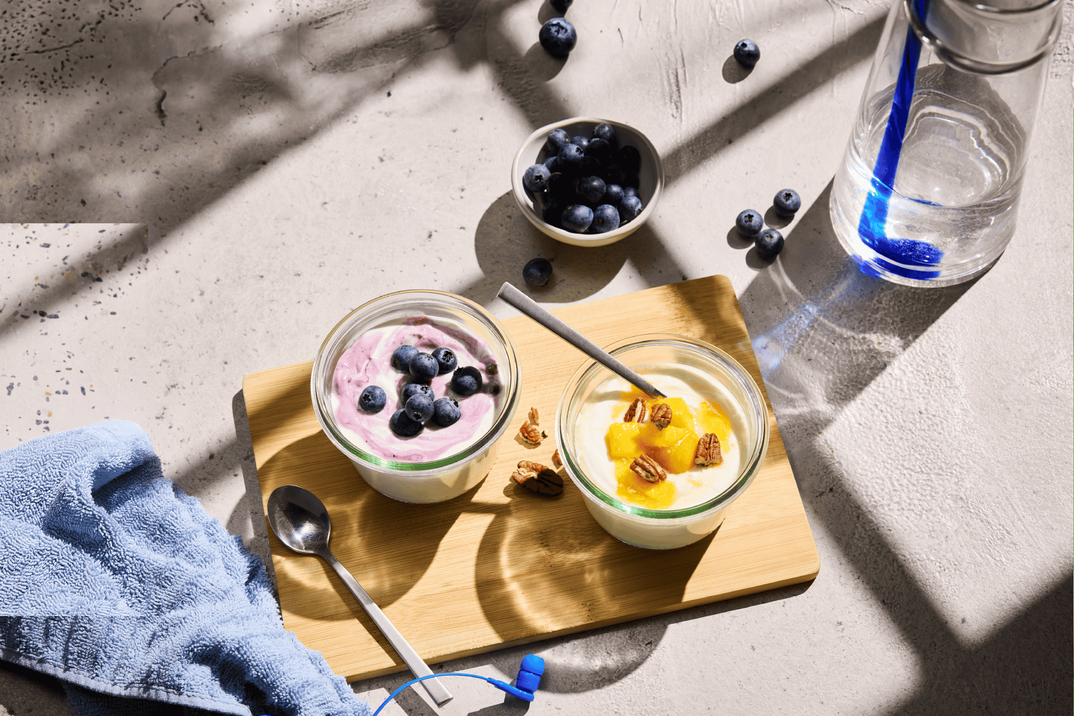 To glas yoghurt med blåbær og fersken, en vandflaske og et håndklæde på et bord.