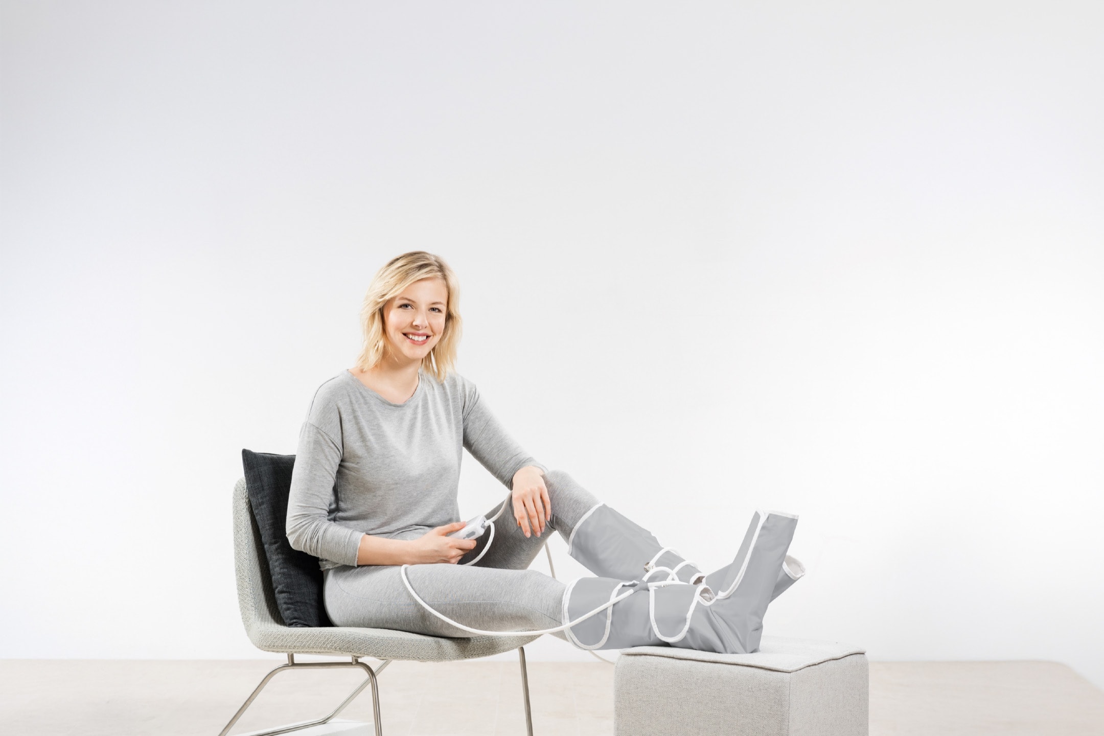 Kvinde i grå loungewear med benmassageapparat og kontrolenhed.
