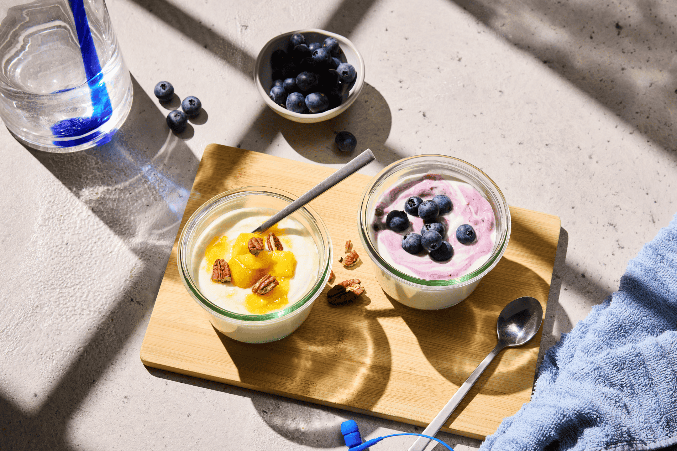 To skåle yoghurt med frugt og nødder på et træbræt, med blåbær og vand.