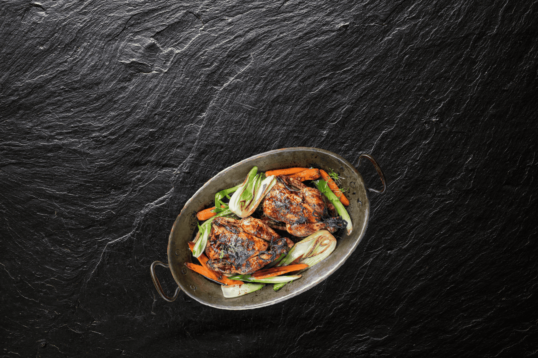 Helstegt kylling med gulerødder og fennikel i en oval pande på en mørk stenoverflade.