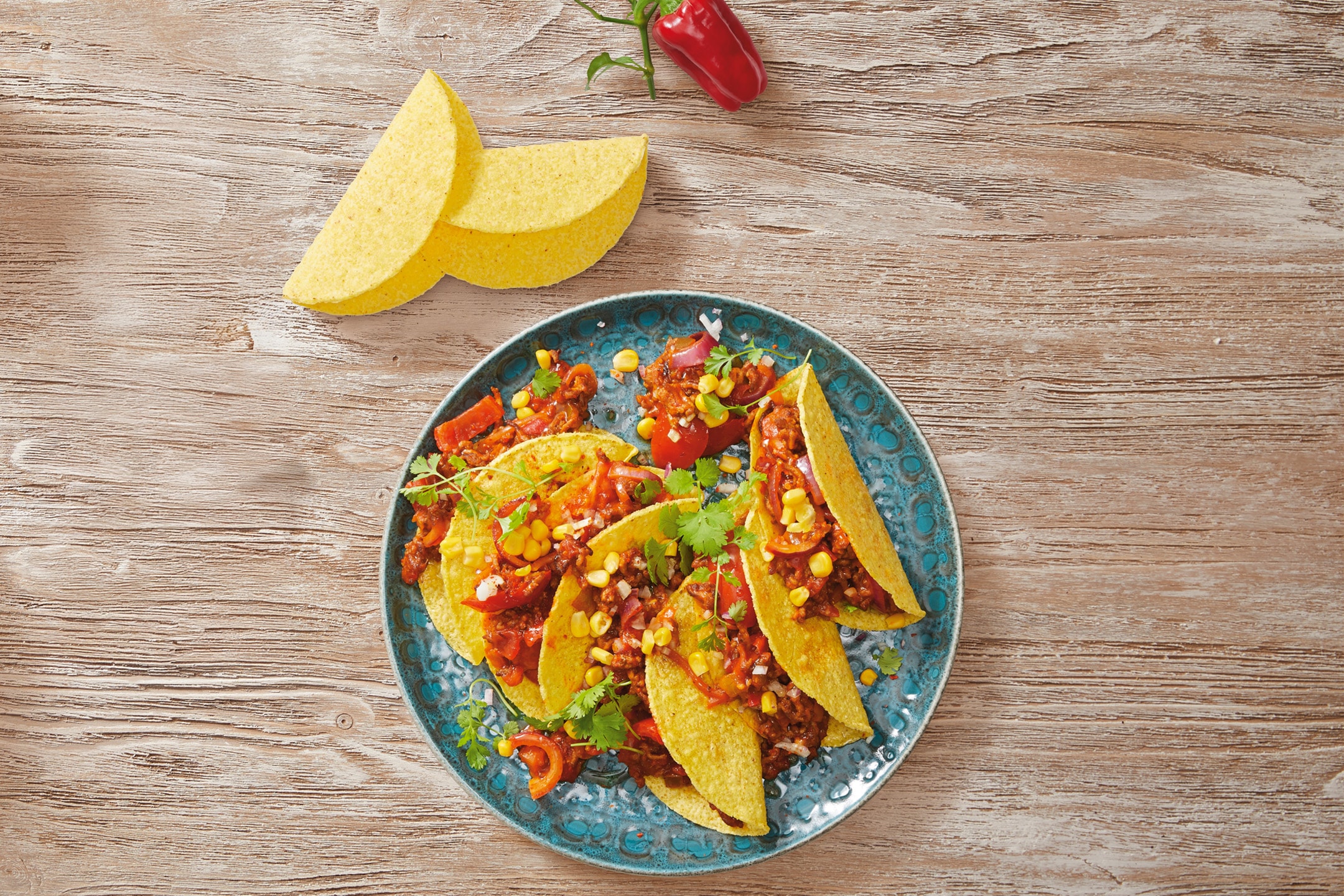 Tacoskaller fyldt med hakket kød, majs, peberfrugt og løg på et blåt fad.