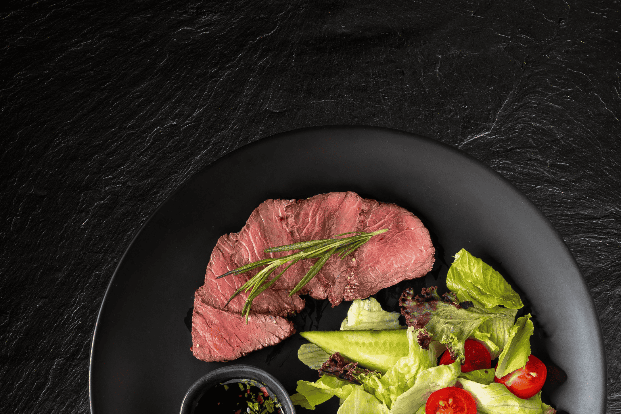 Skiver af oksekød med rosmarin, salat og dressing på en sort tallerken.