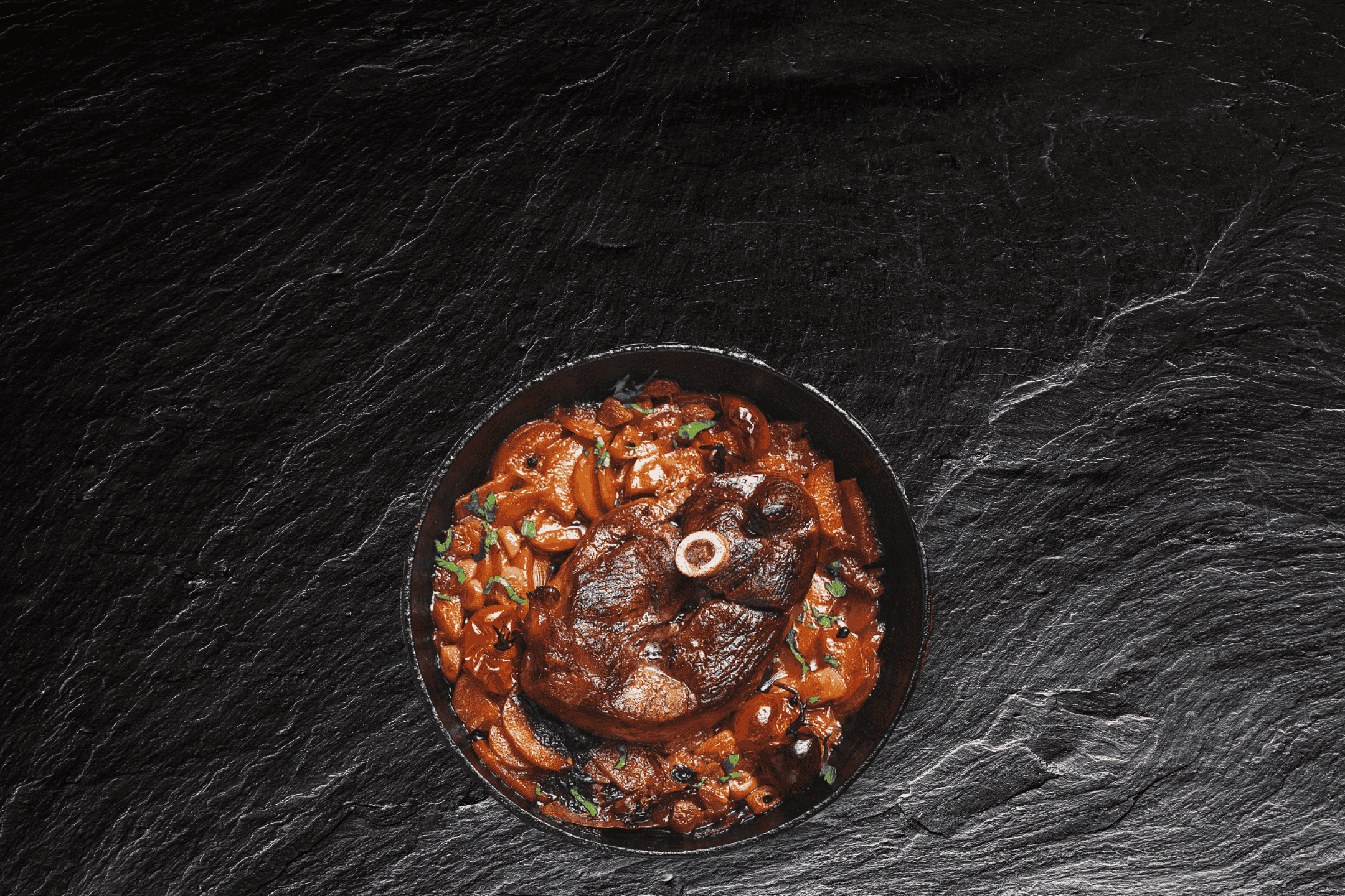 Langtidsstegt lammekølle med grøntsager i en sort gryde på en mørk skiferbaggrund.