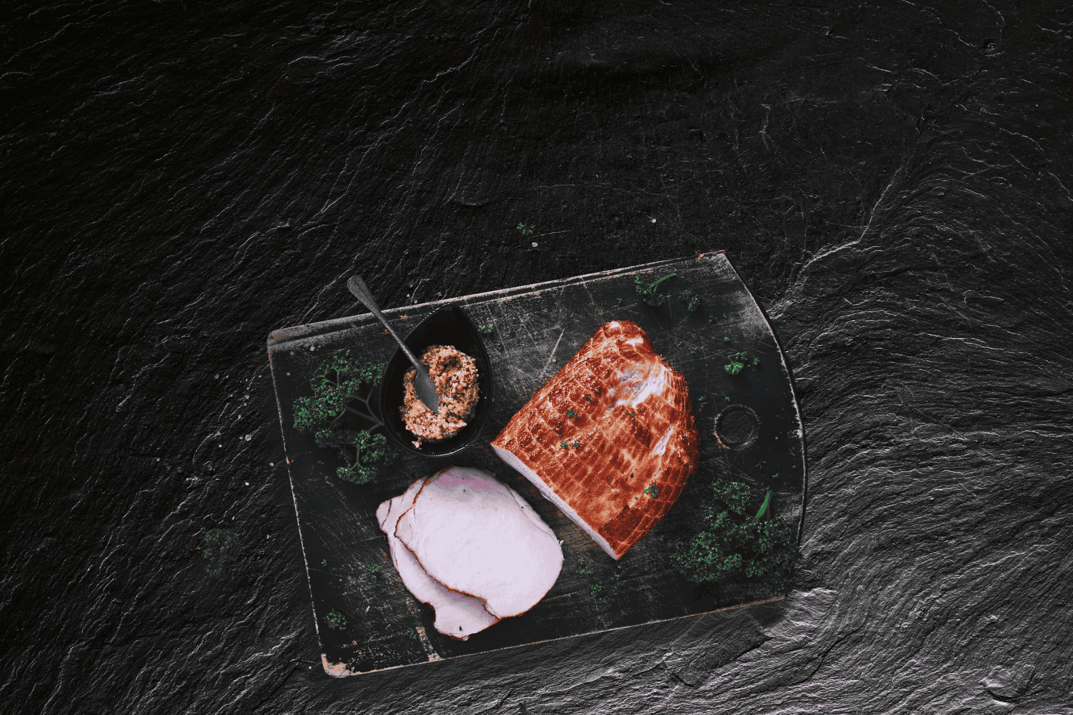 Skiver af stegt svinekød med sennep og persille på et mørkt skærebræt.