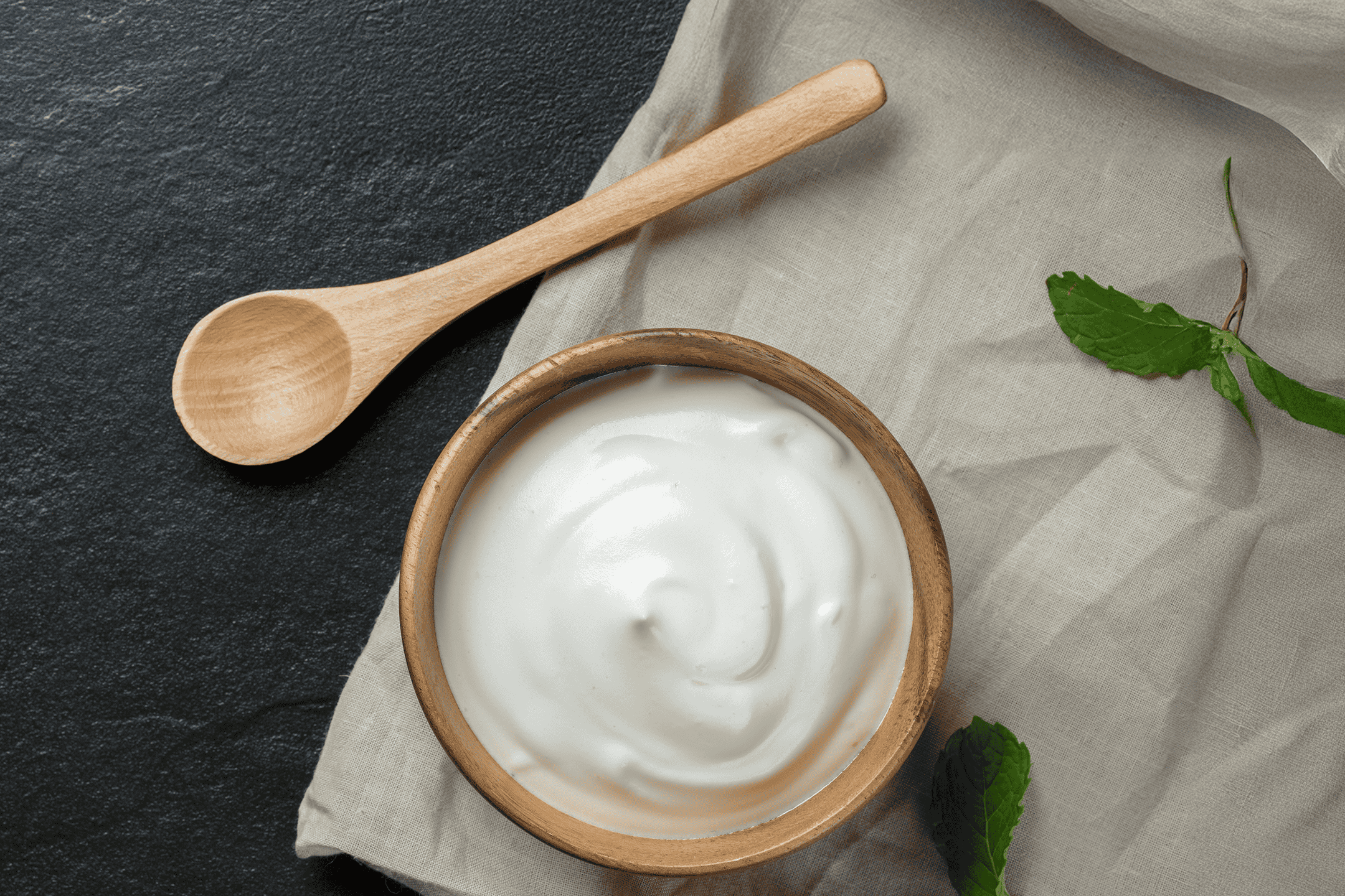 Græsk yoghurt i træskål med træske og mynteblade på mørk baggrund.
