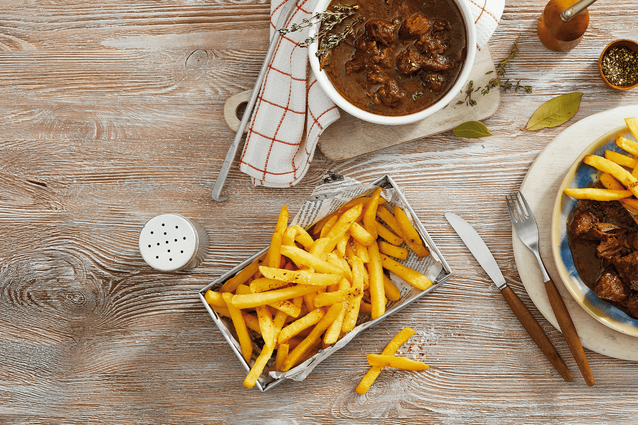 Gryderet med pommes frites, krydderier og bestik på et træbord.