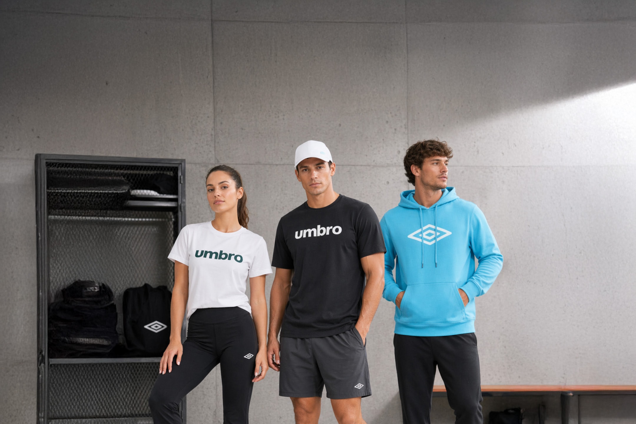 Tre personer i sportstøj, herunder t-shirts, shorts, leggings og en hættetrøje, står i et omklædningsrum.
