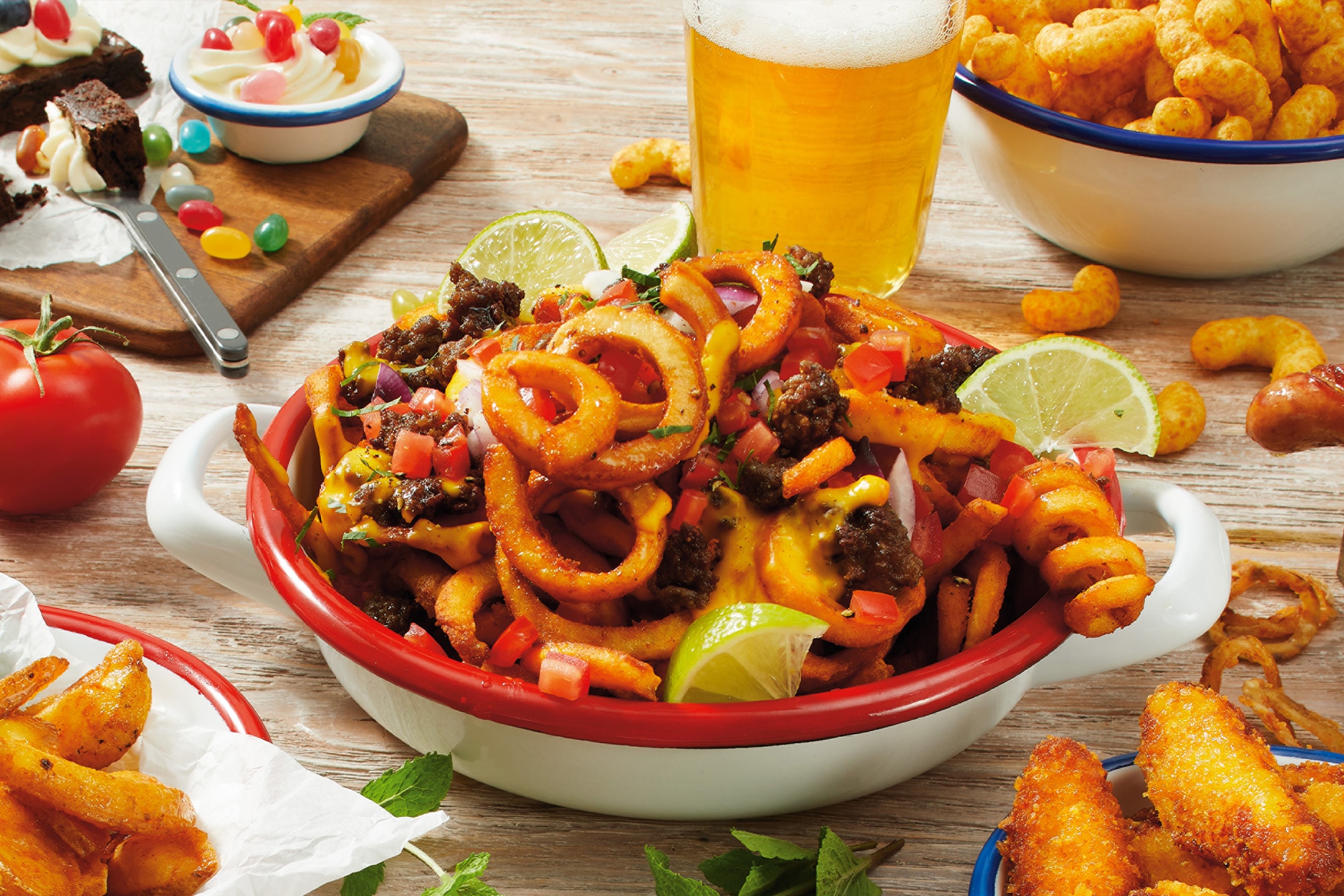 En skål med krøllede fritter toppet med kød, ost og grøntsager, omgivet af snacks og dessert.