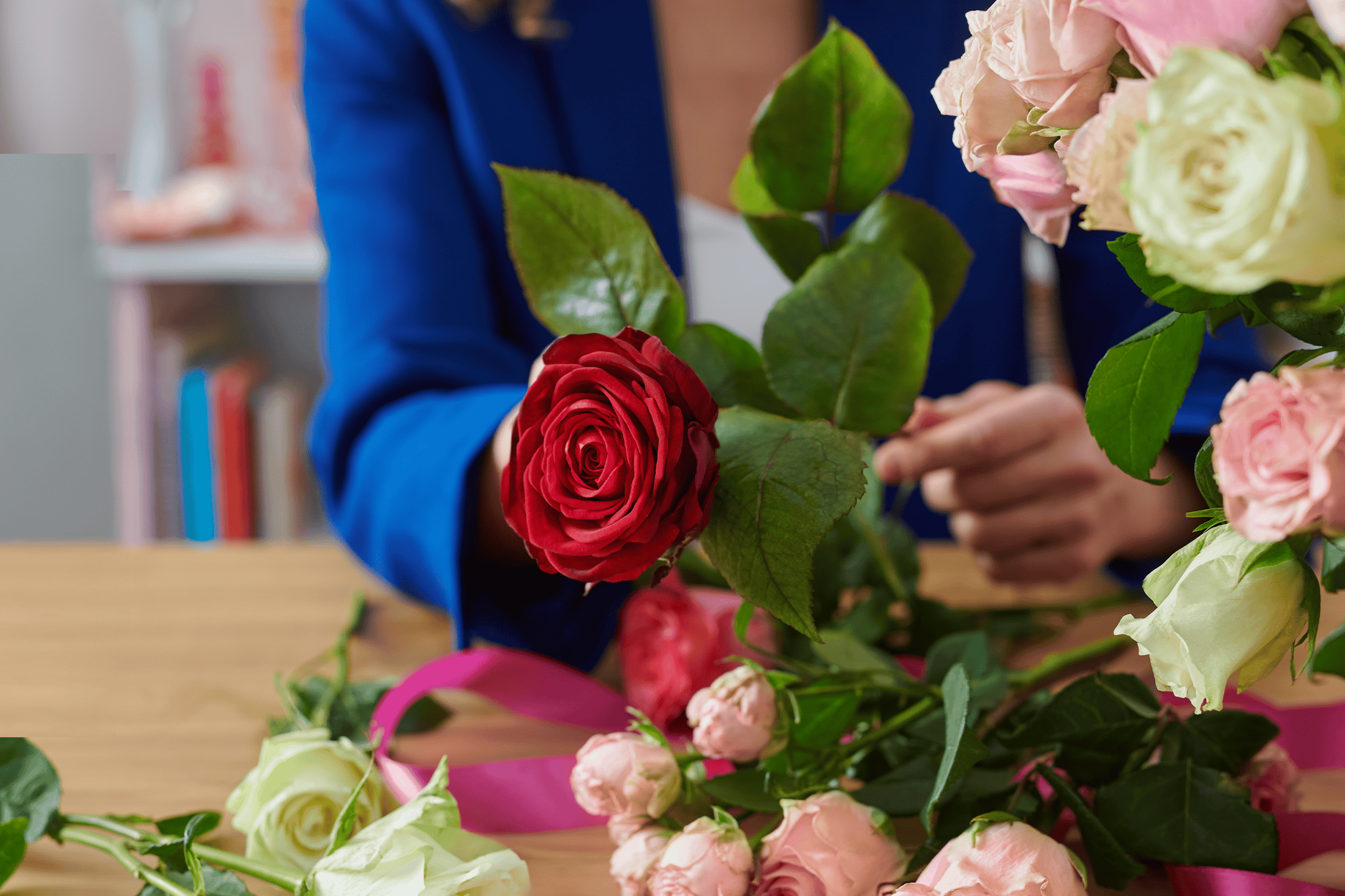 En person i en blå jakke arrangerer røde og lyserøde roser på et træbord.