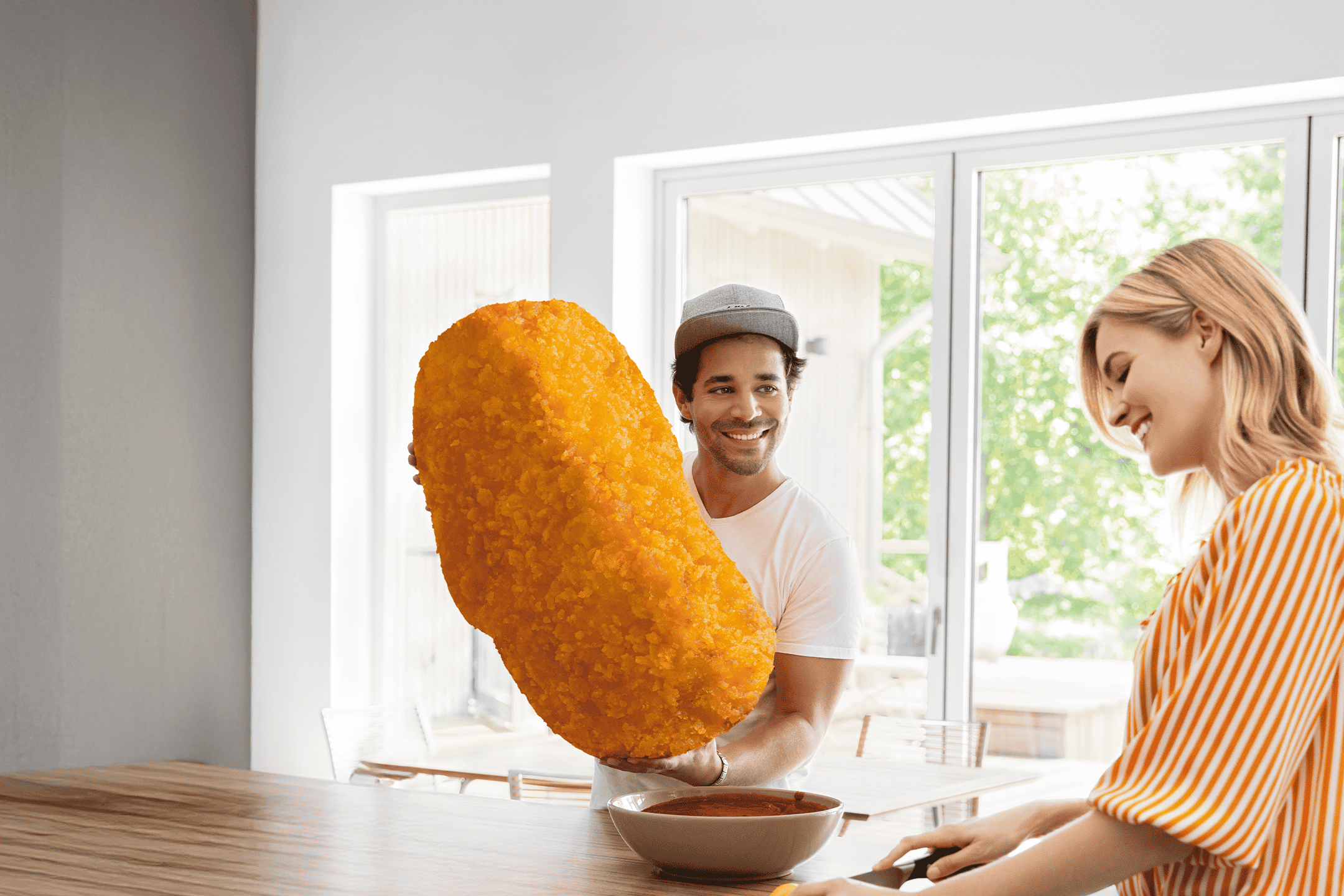 Mand holder kæmpe friturestegt madvare, kvinde smiler ved køkkenbordet.