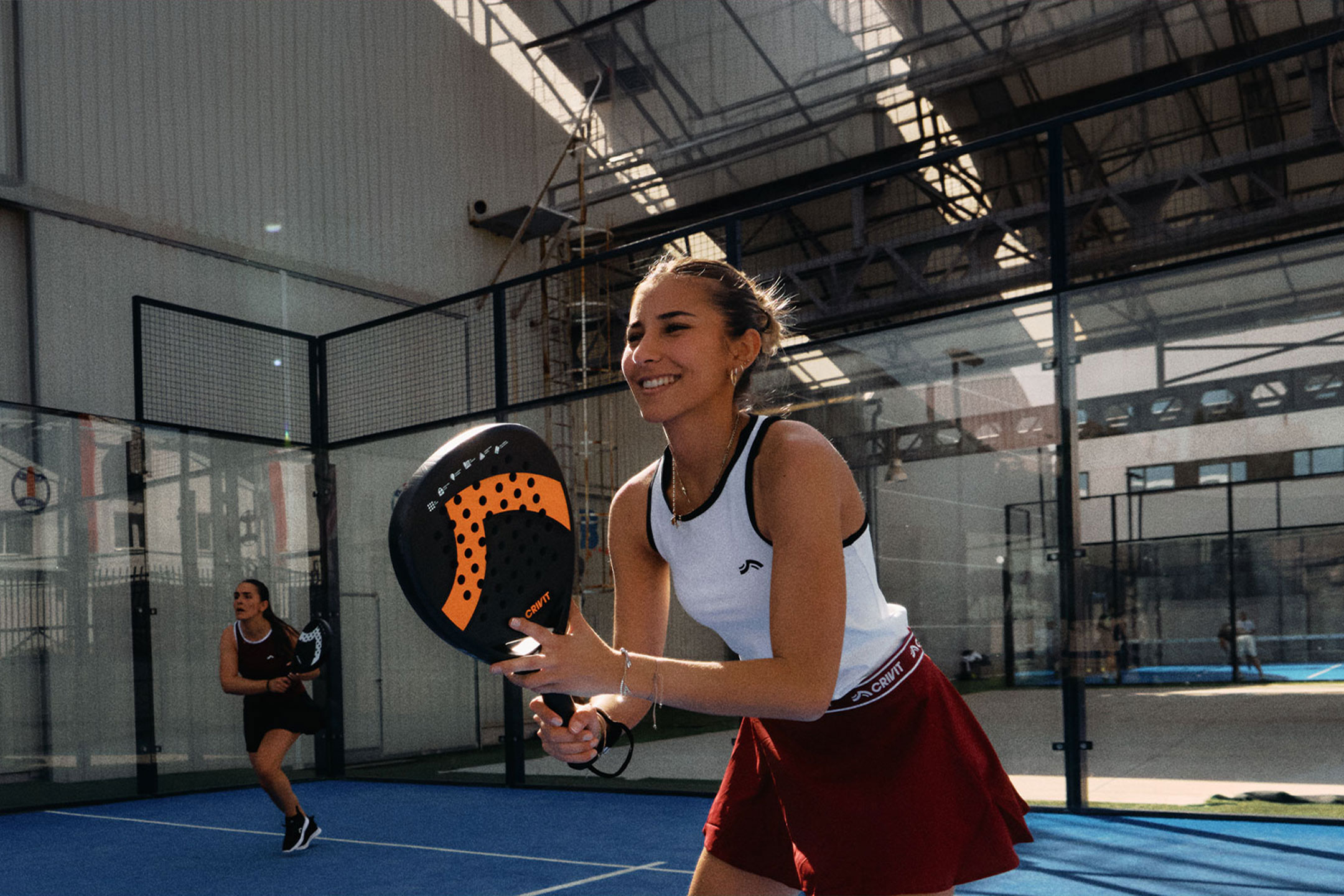 aiLabeling.aiLabelForImageAltText: To kvinder spiller padeltennis, den ene smiler og holder en ketcher, på en blå bane.