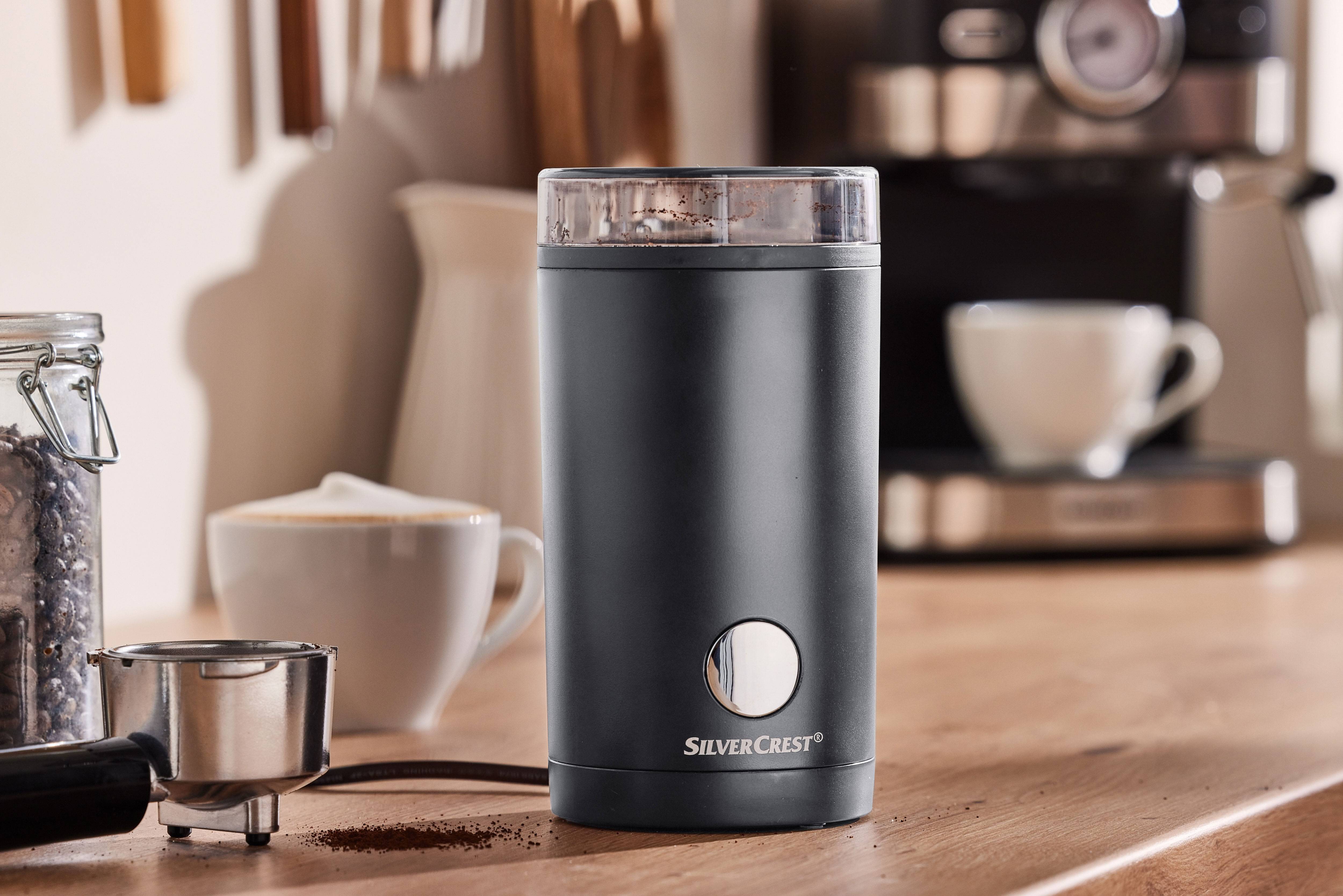 Sort kaffekværn med malet kaffe, kaffemaskine og kopper på et træbord.