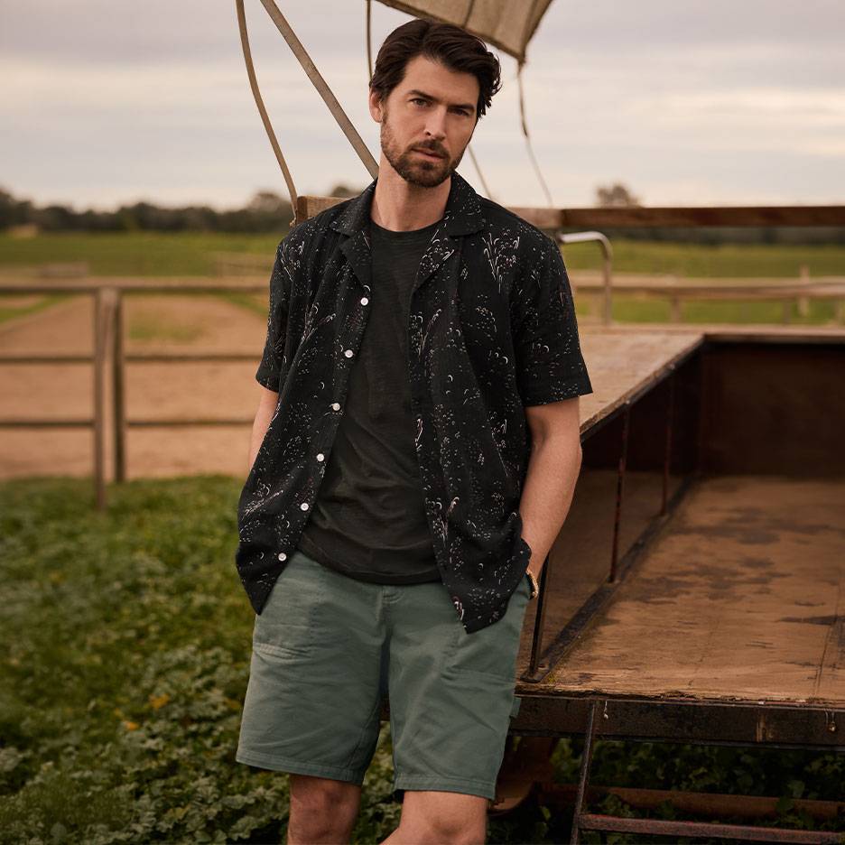 Mand i sort mønstret skjorte, T-shirt og grønne shorts, stående ved en trailer.