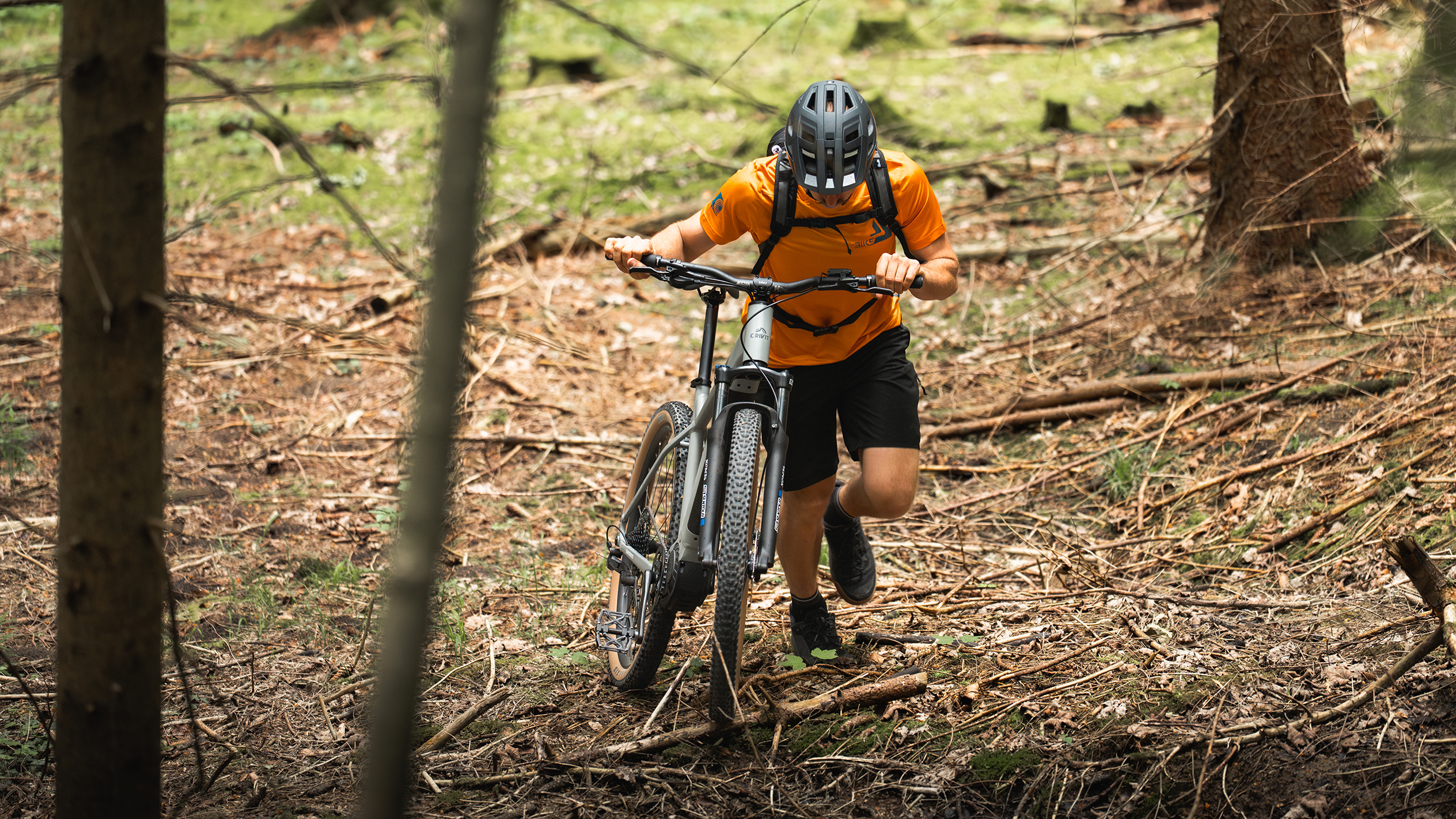 En cyklist i orange skjorte skubber en mountainbike elcykel gennem en skov.