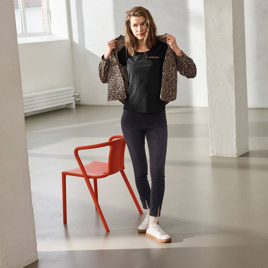 Kvinde i sort t-shirt med tekst, jeans og leopardprint jakke, stående ved siden af en rød stol.