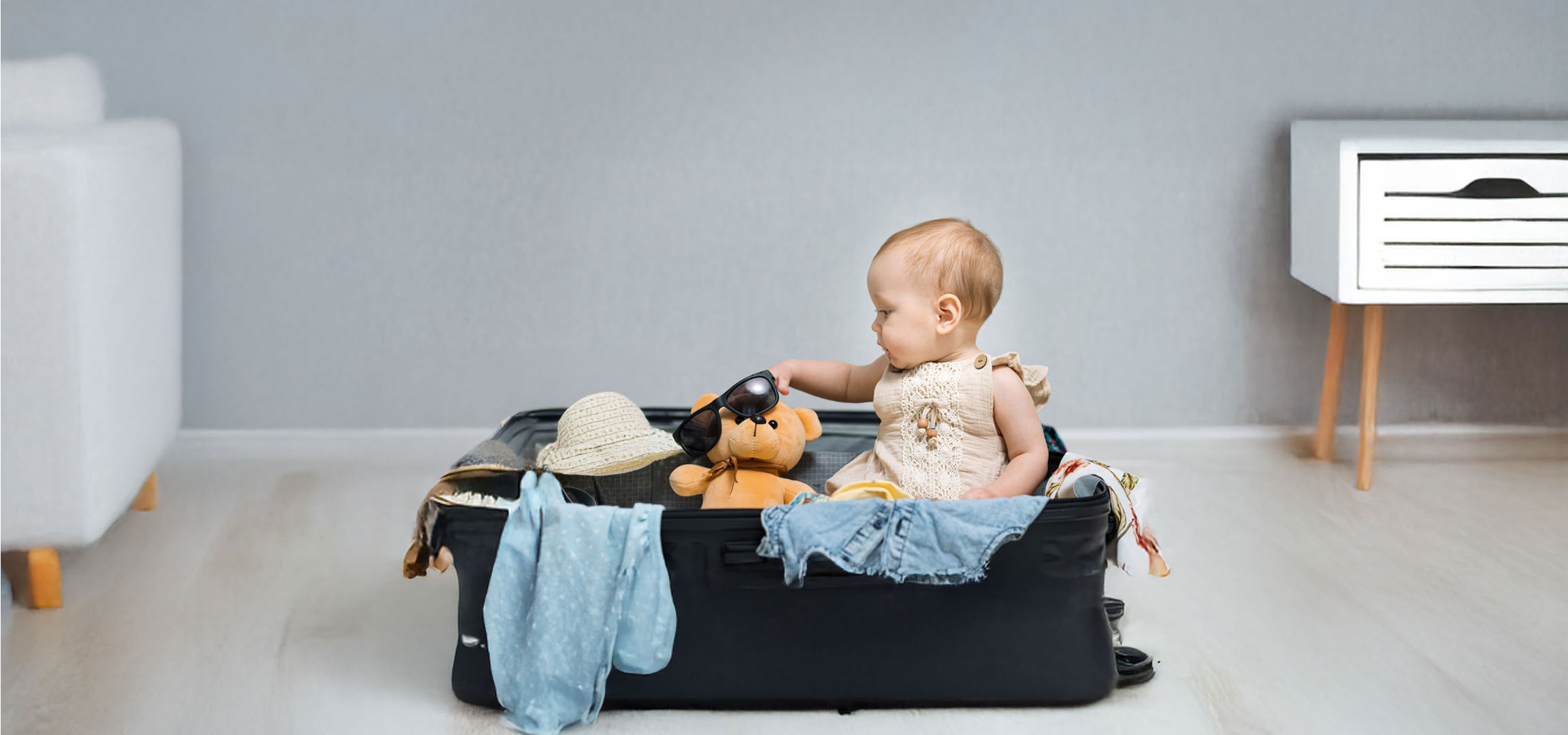 Baby pakker tøj og en bamse i en kuffert.