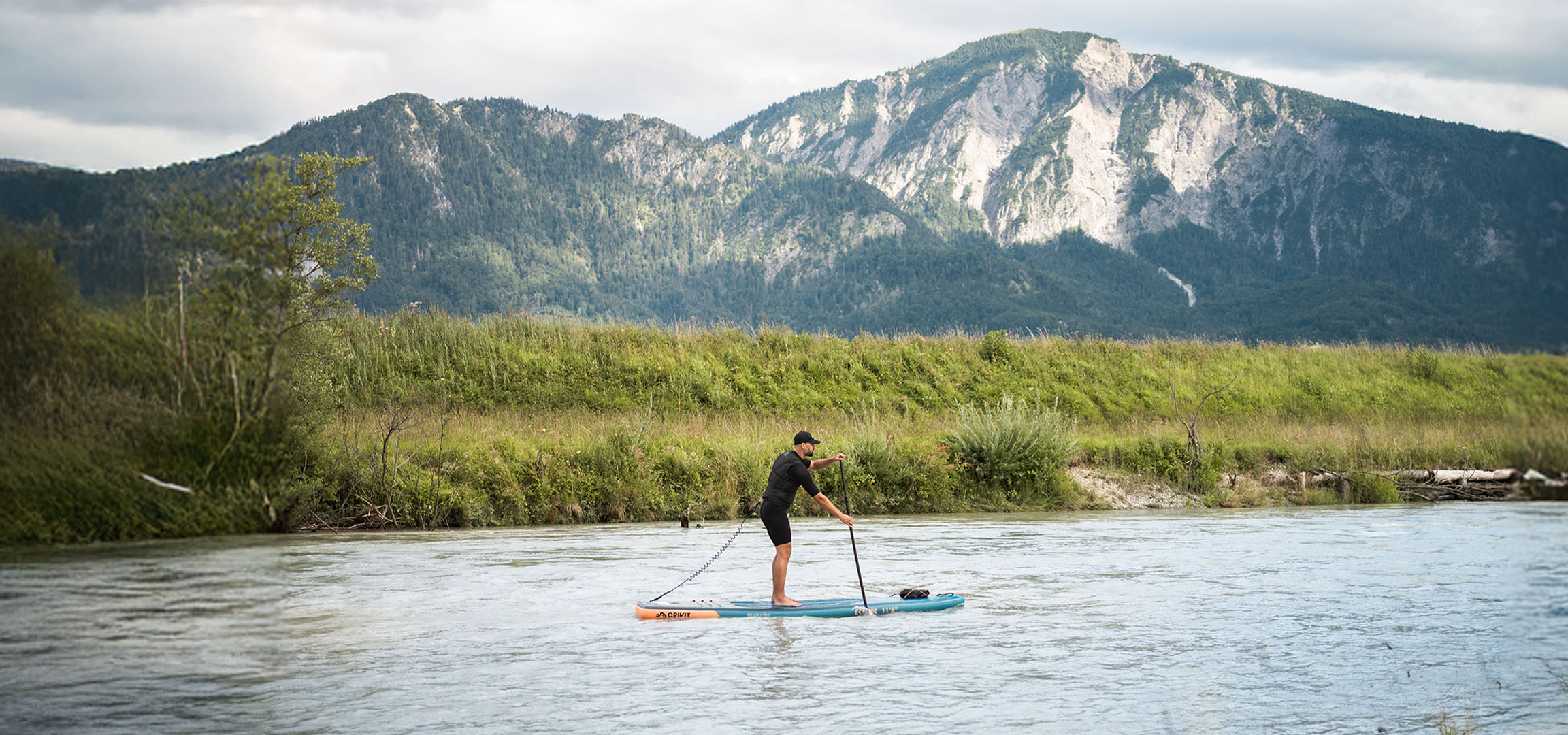 Mand på paddleboard på en flod med bjerge og frodig vegetation i baggrunden.