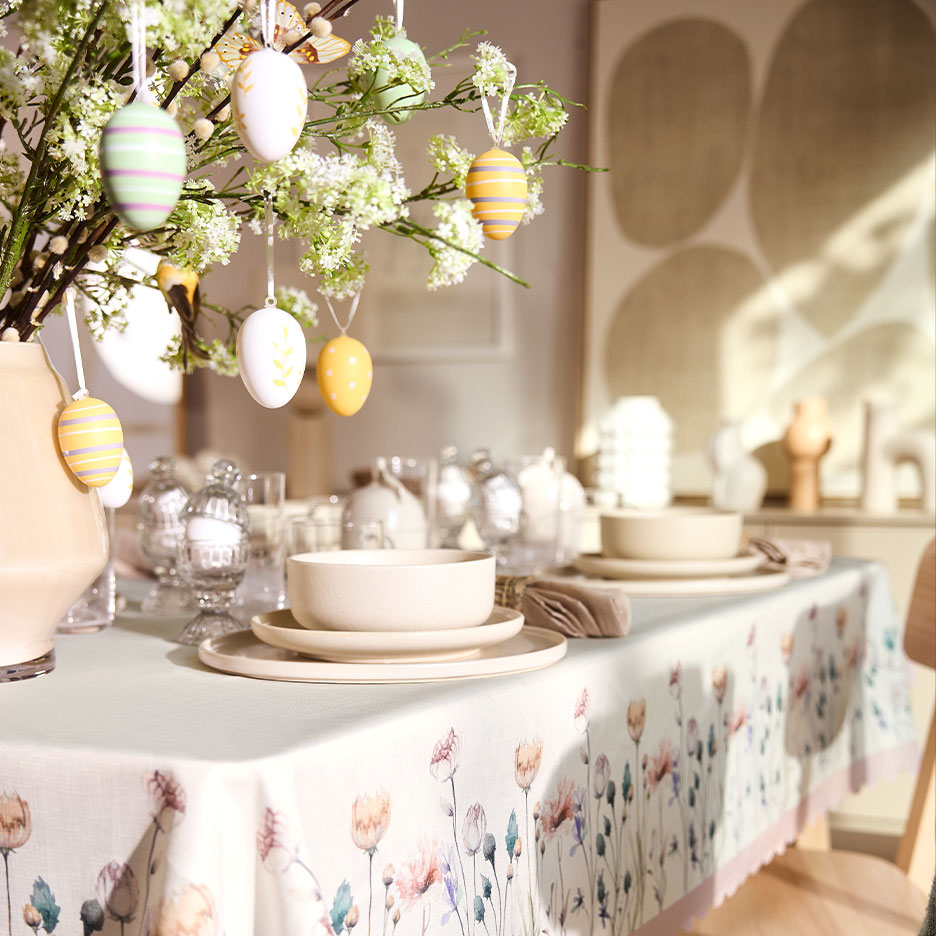 Påskebord dækket med blomstret dug, keramisk service og grene dekoreret med æg.