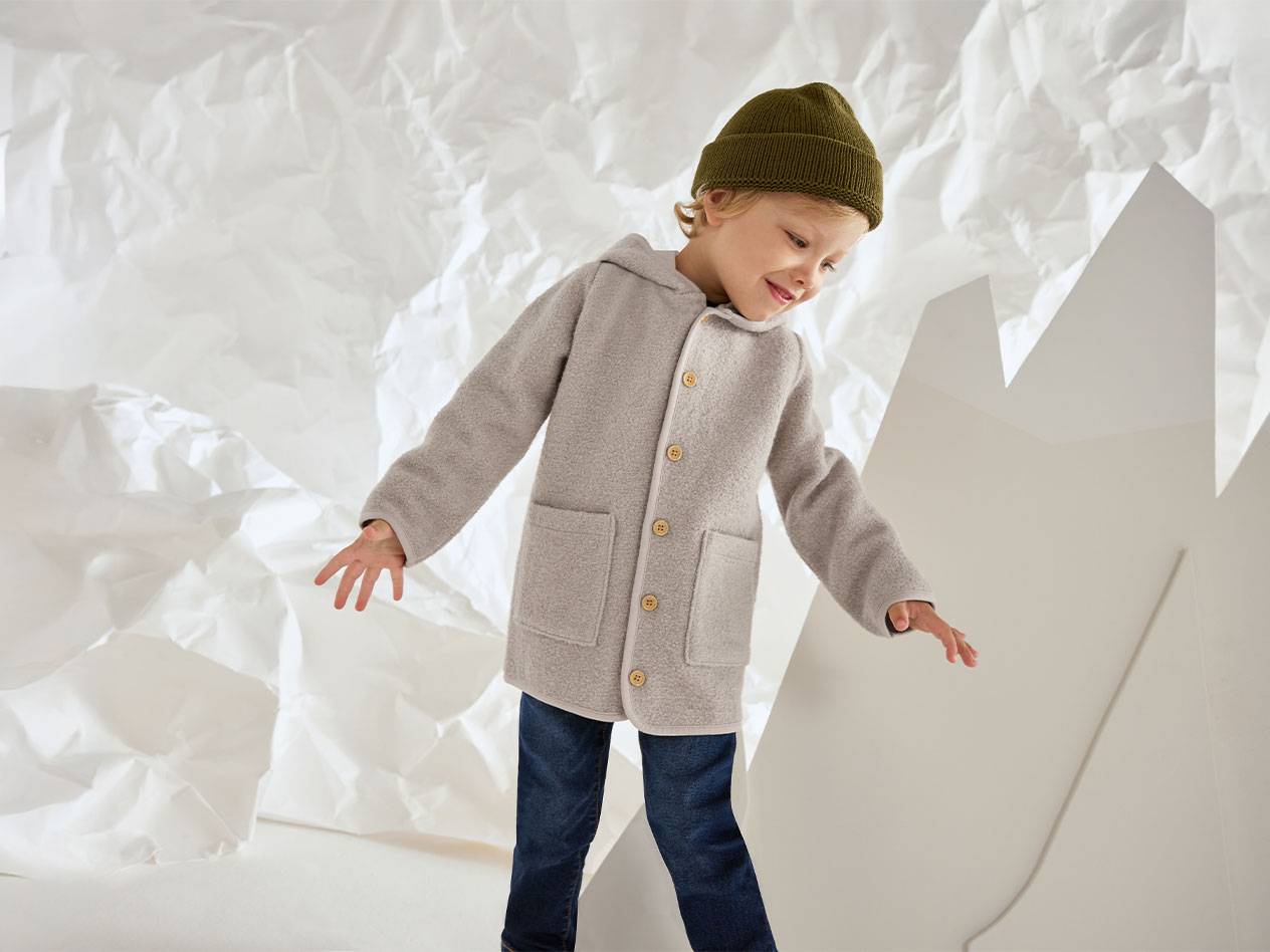 Dreng i grå uldjakke og grøn hue, smilende mod en baggrund af krøllet papir.