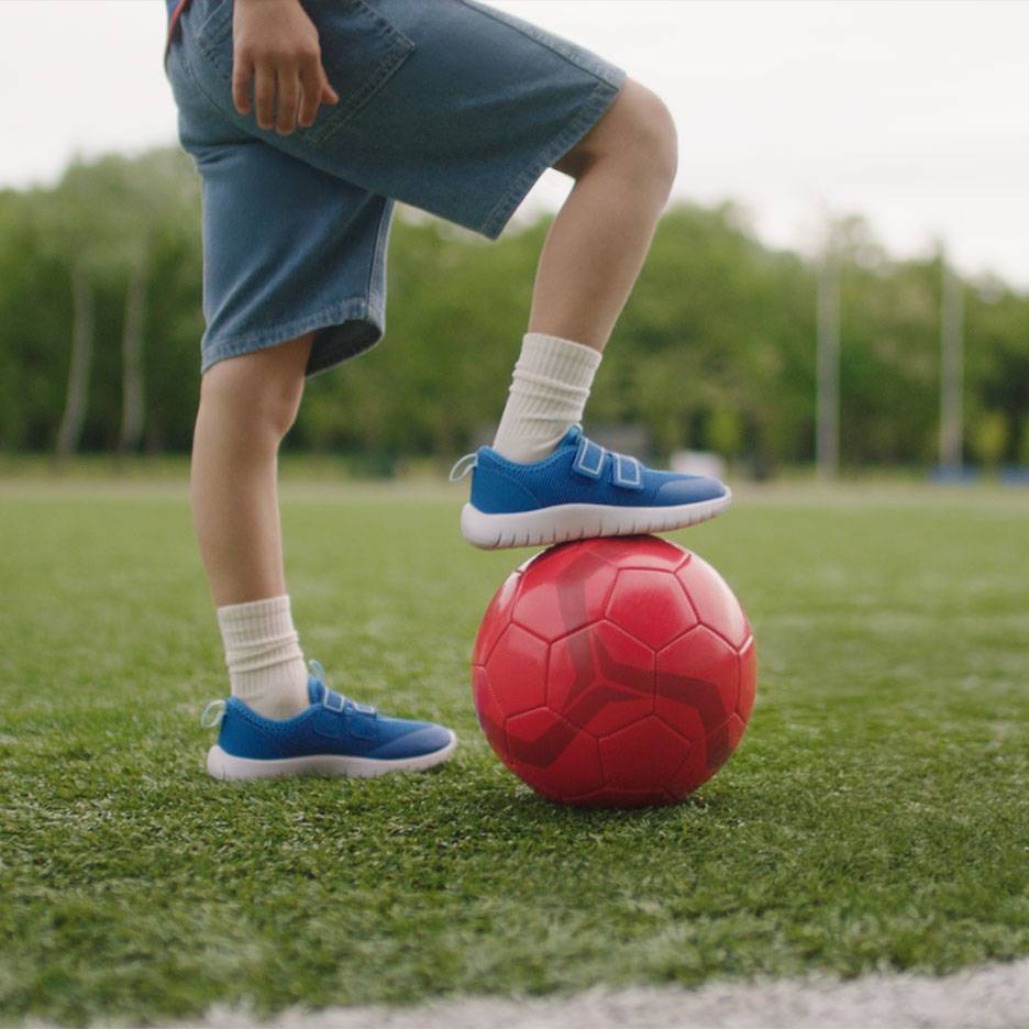 Barn i shorts og blå sneakers, med den ene fod på en rød fodbold.