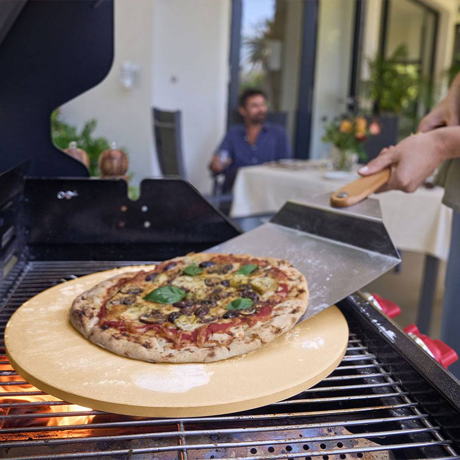 Pizza på en pizzasten på en grill, med en pizzaspade og en mand i baggrunden.