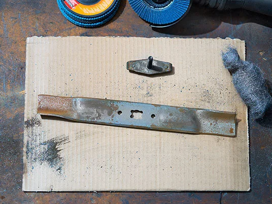 Rusten plæneklipperklinge på pap med slibeskiver og ståluld.