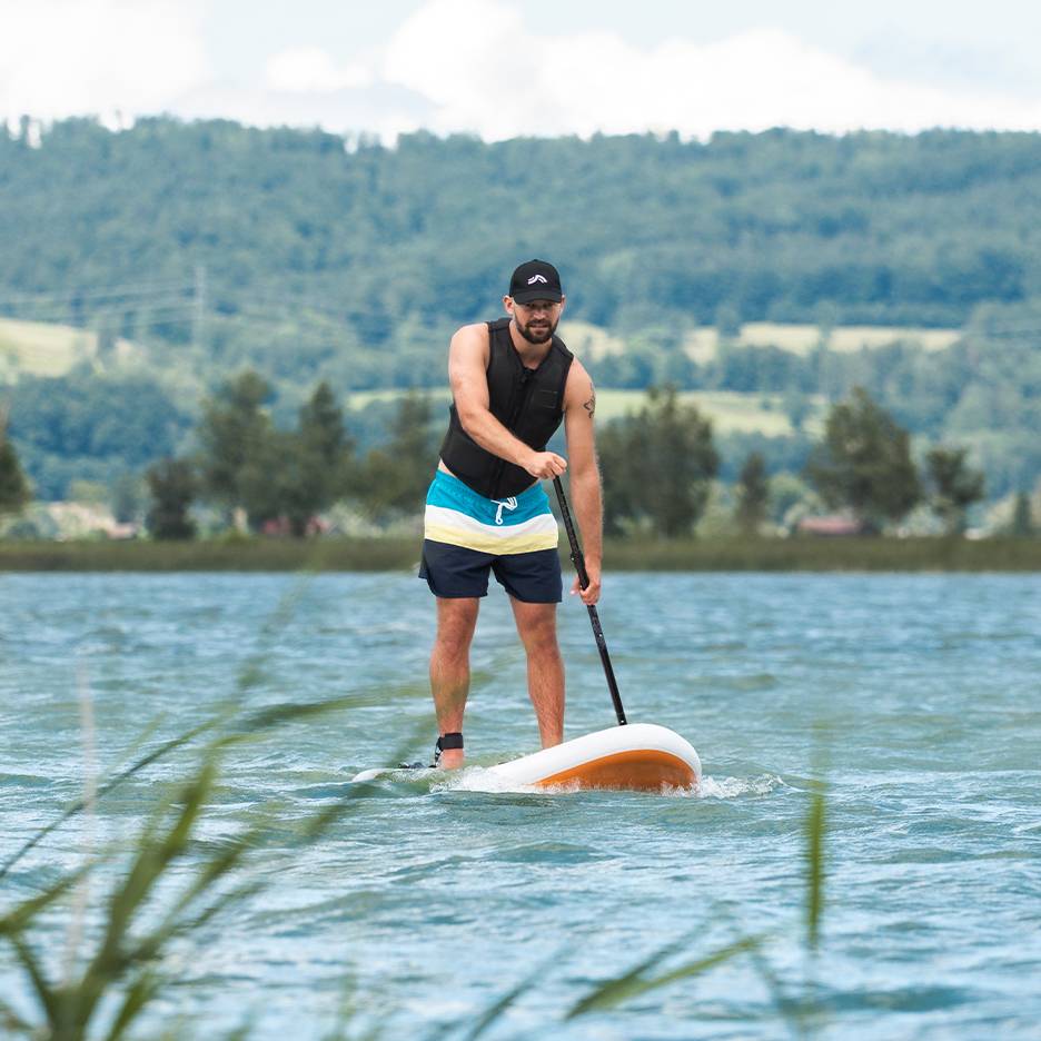 Mand stand-up paddleboarder på en sø, iført redningsvest og badeshorts.