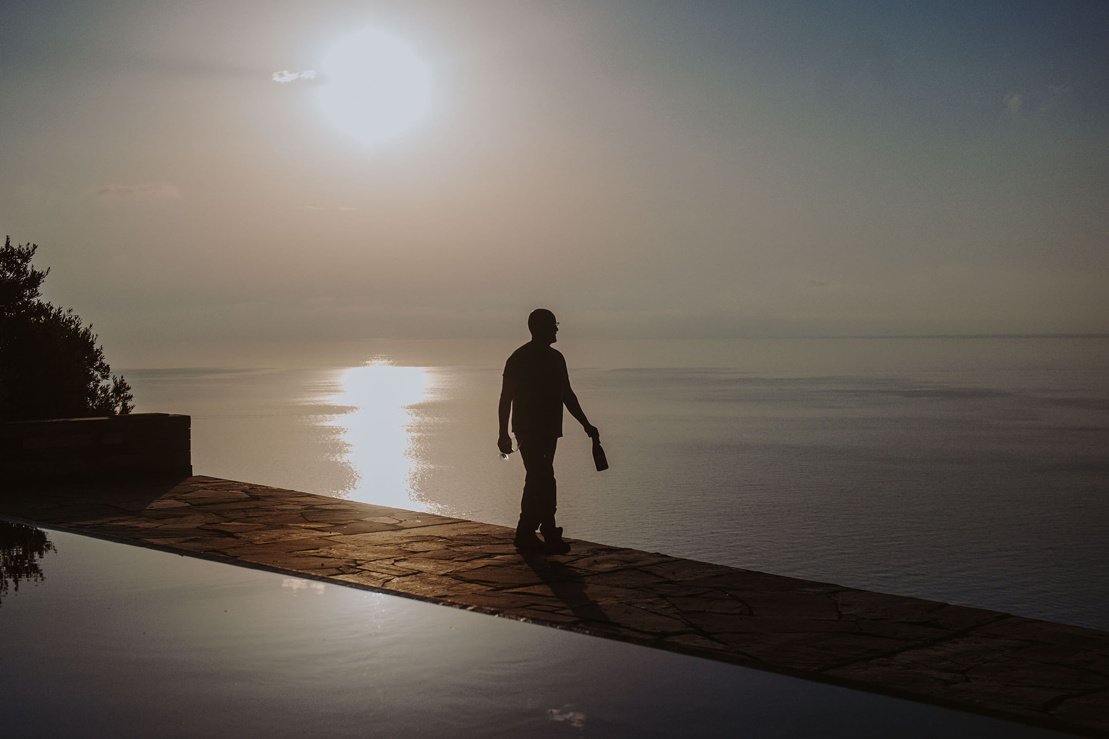 Silhuet af en person med flaske og glas ved en infinitypool ved solnedgang