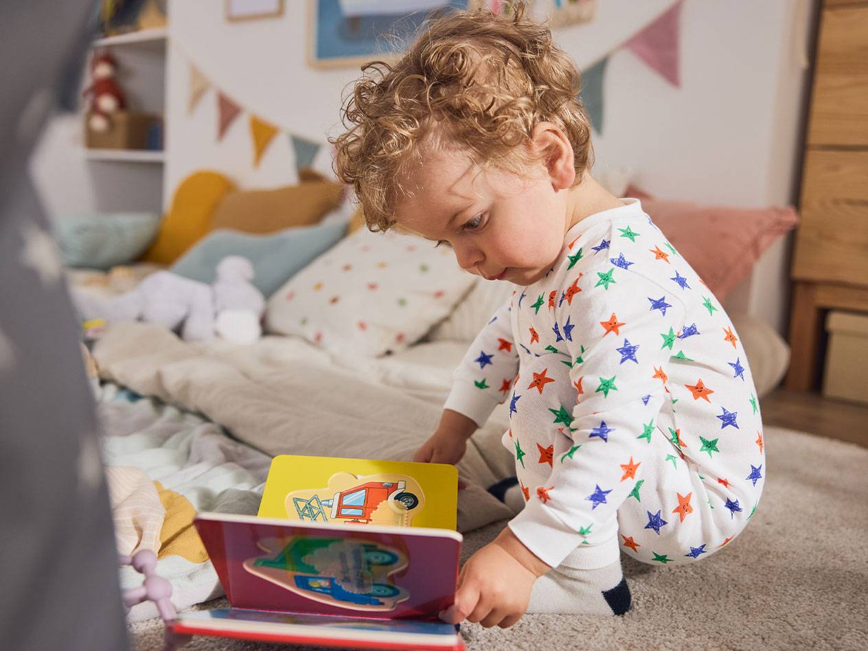 Et lille barn i stjernepyjamas, siddende på tæppet, læser en bog.