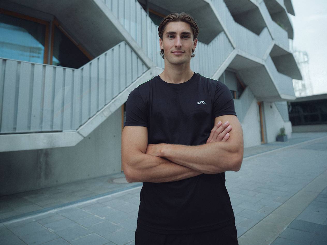 Mand i sort t-shirt med logo og sorte shorts, står udendørs med krydsede arme.