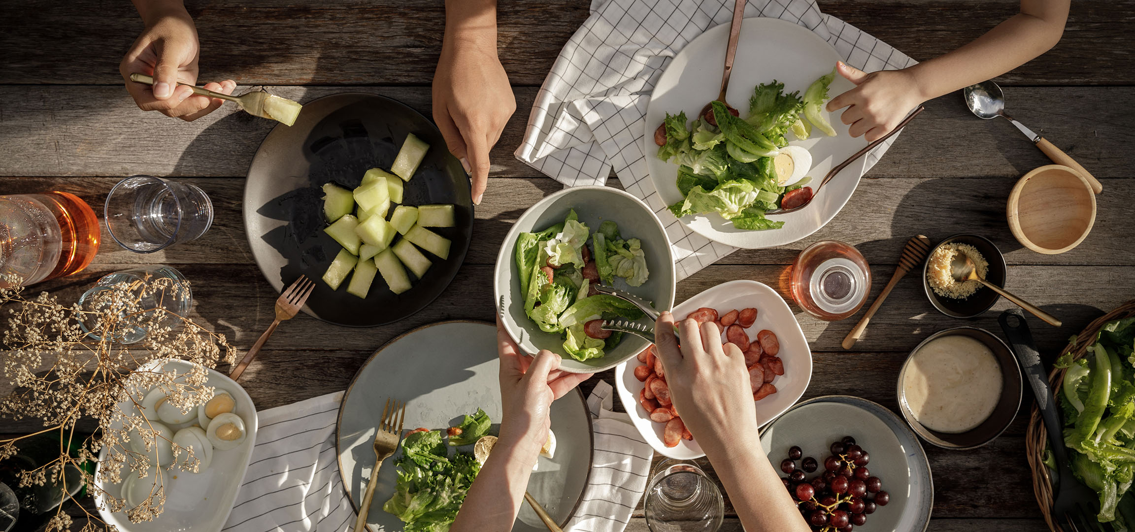 Oversigt over et bord fyldt med salat, pølser og melon, med folk der serverer mad.
