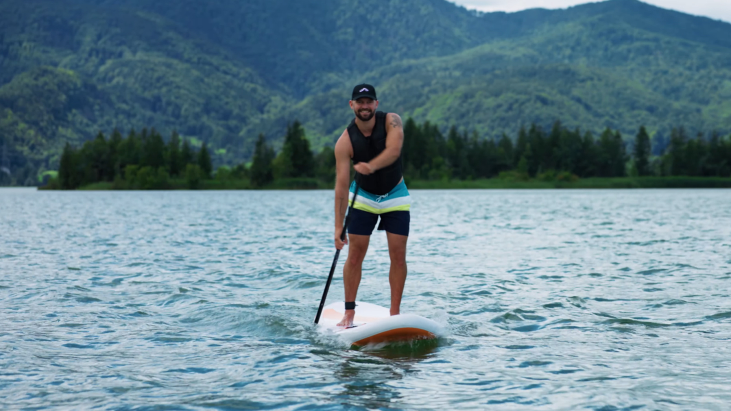 Mand på paddleboard på en sø med bjerge i baggrunden.
