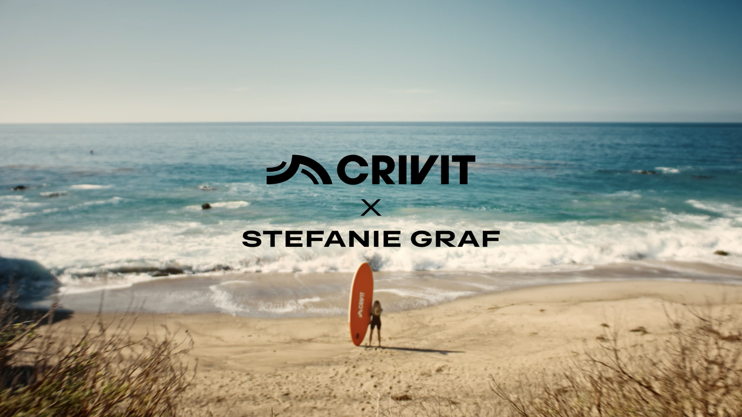 Kvinde med et orange SUP-board på en sandstrand, med havet i baggrunden.