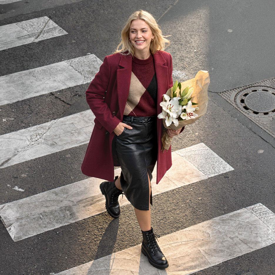 Smilende kvinde i bordeaux frakke, argyle sweater og lædernederdel, krydser gaden med en buket liljer.