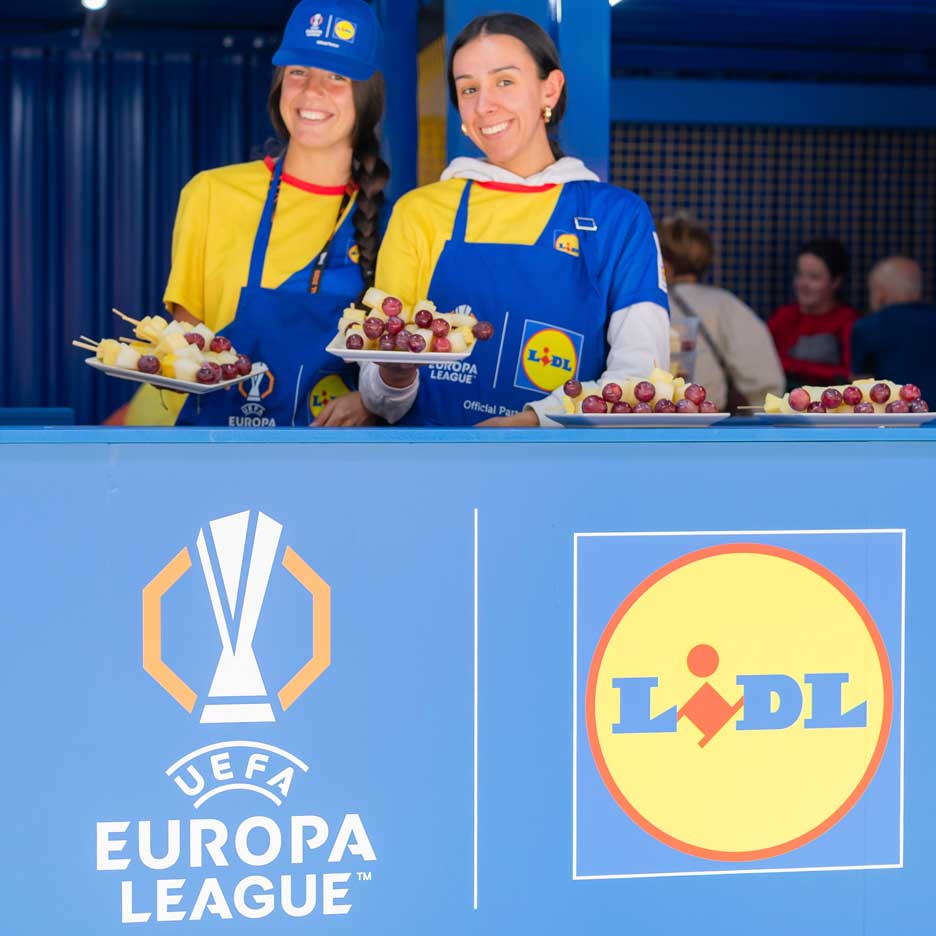To smilende kvinder i Lidl-forklæder tilbyder frugt på spyd ved en UEFA Europa League-begivenhed.