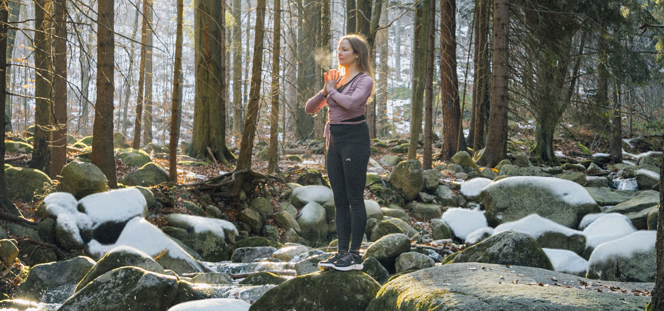 Kvinde i sportstøj laver yoga i en sneklædt skov med en bæk.