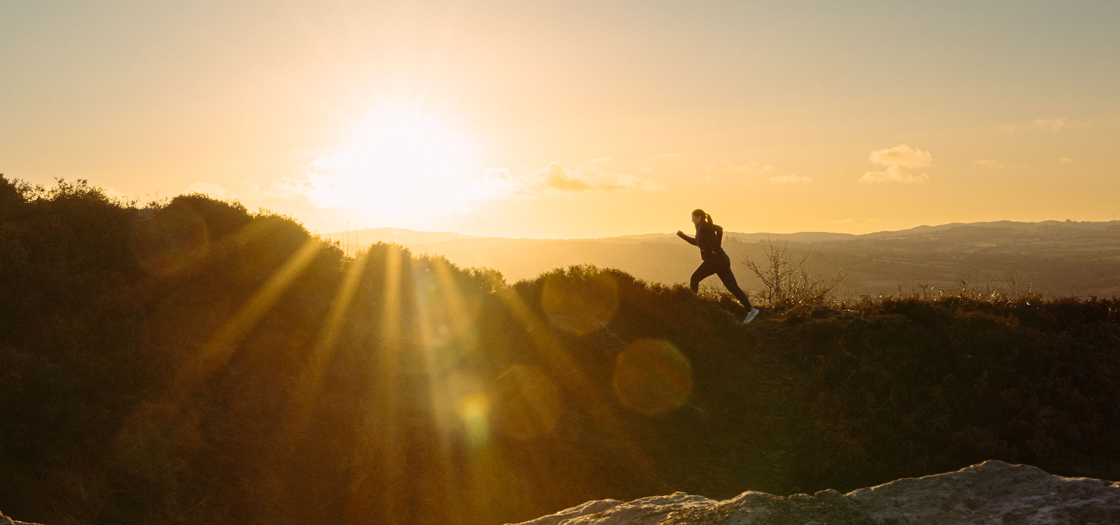 Kvinde løber på en bakke ved solnedgang, med solstråler og et naturskønt landskab.