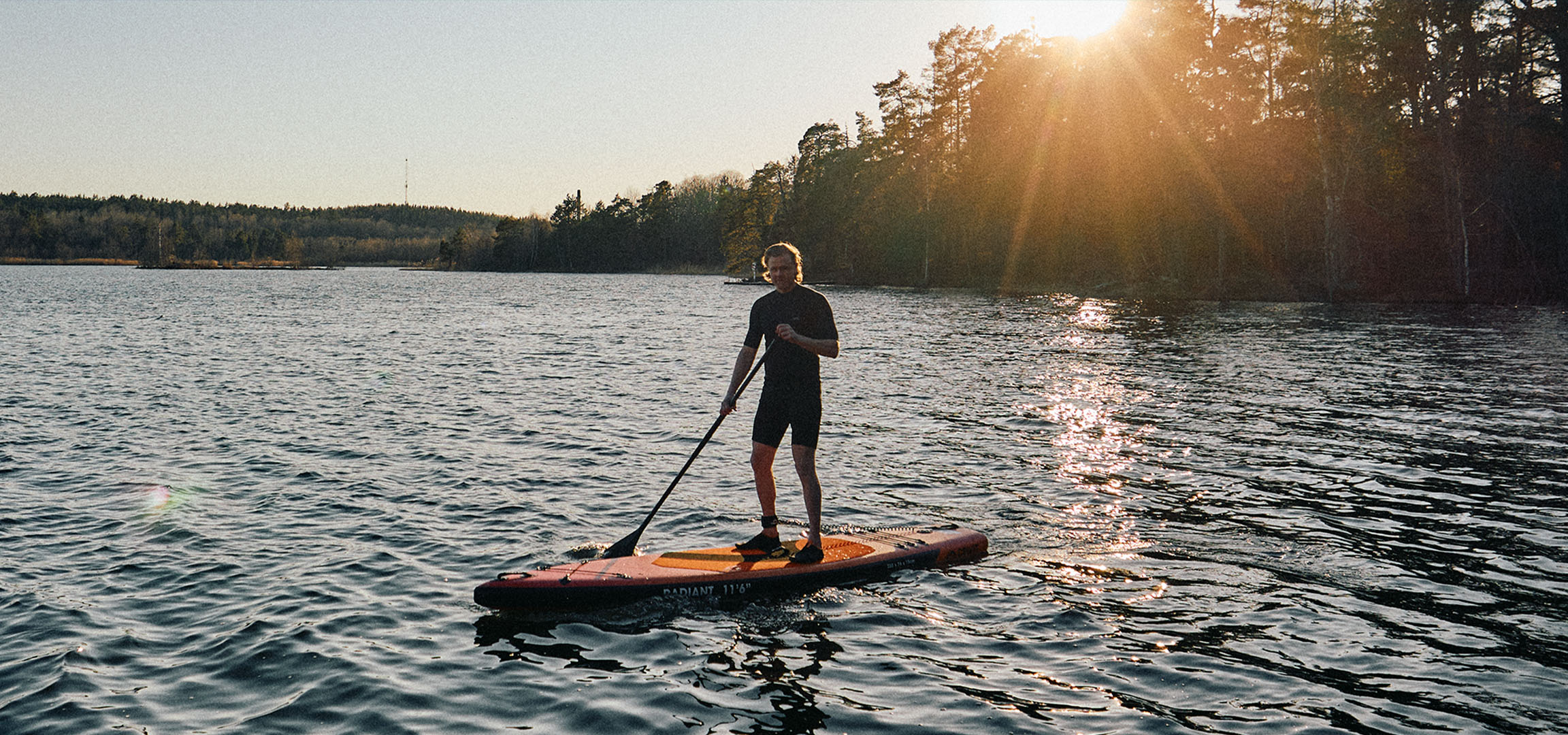 Mand på et stand-up paddleboard på en sø ved solnedgang.