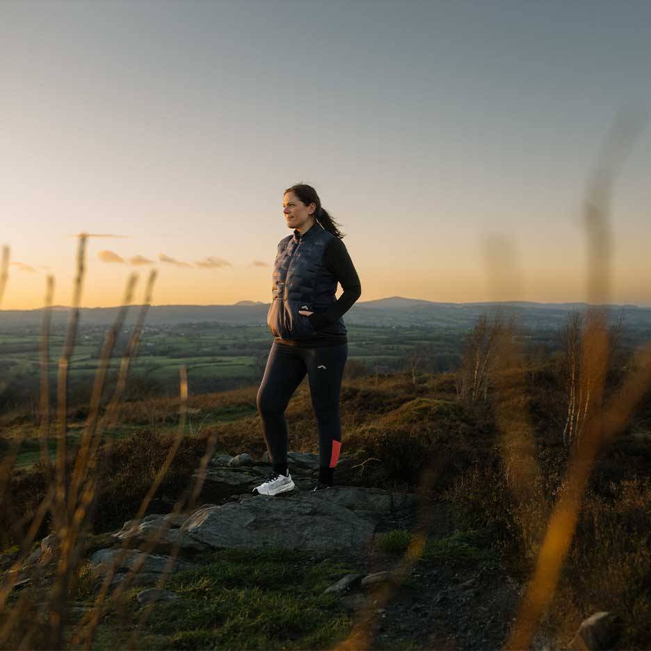 Kvinde i vatteret vest og leggings står på en bakketop ved solnedgang.