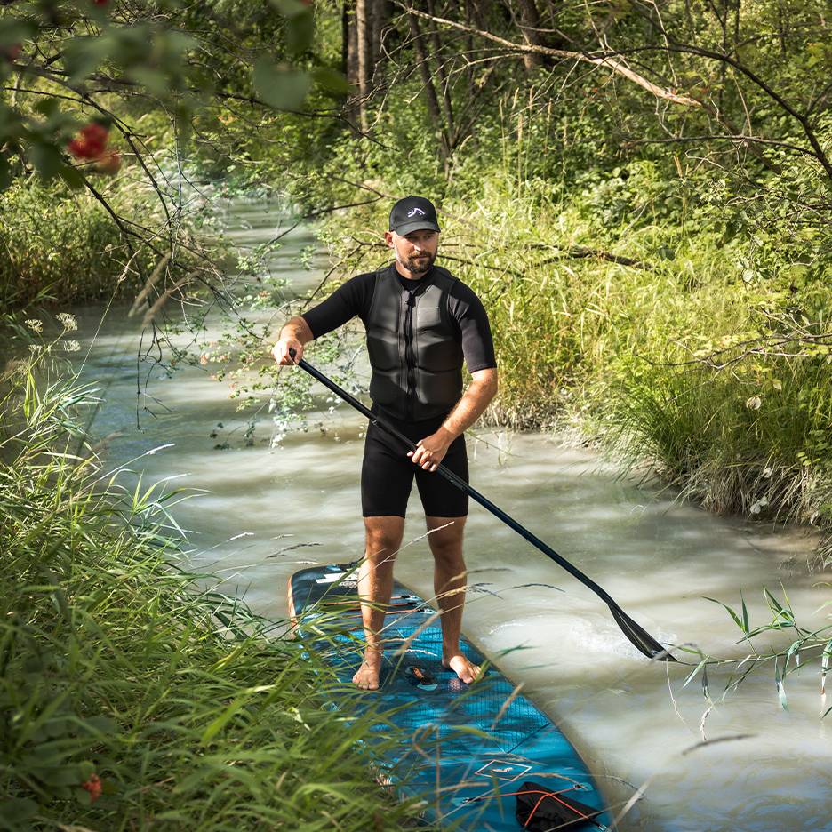 Mand i våddragt og redningsvest står på et paddleboard i en bæk.