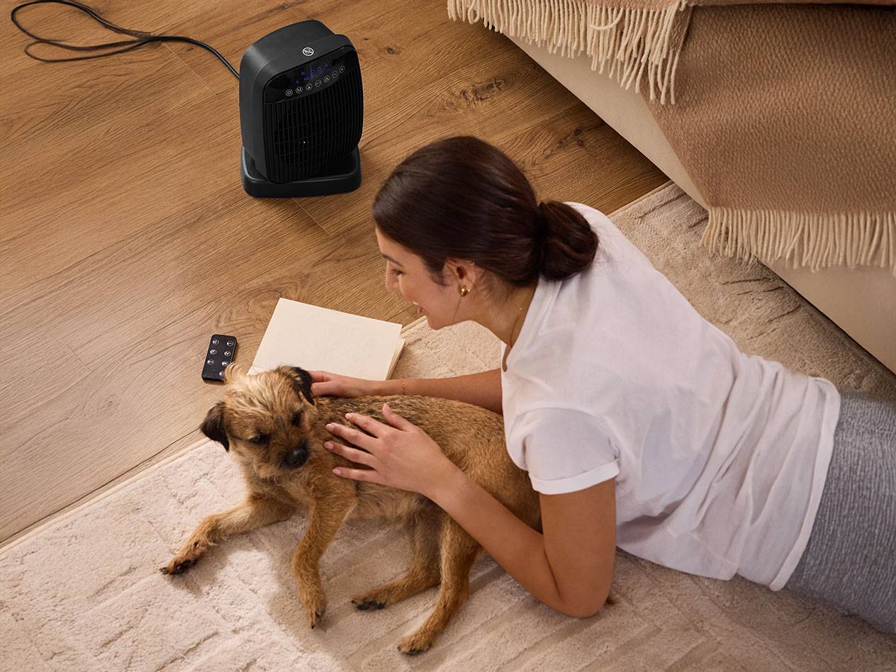 Kvinde kæler med en hund ved siden af en sort rumvarmer på gulvet.