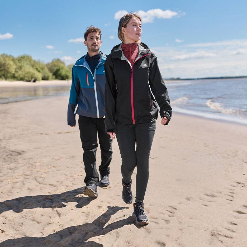 Par i vandrejakker og bukser går på en sandstrand ved havet.