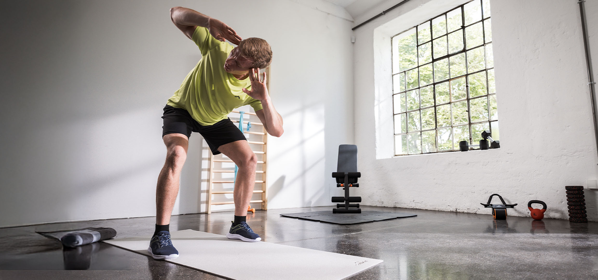 Mand træner i et fitnesscenter, bruger en yogamåtte og håndvægte.