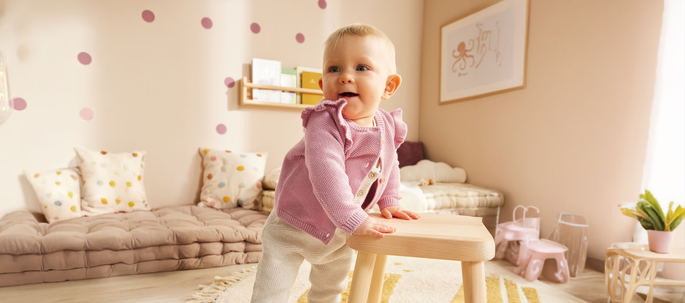 En baby i en sød lyserød cardigan står ved en træskammel på sit børneværelse.