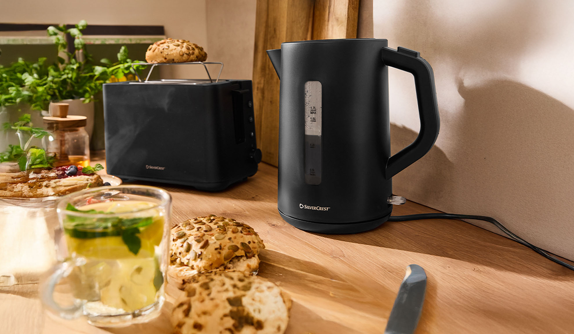 Sort SilverCrest elkedel og brødrister på et træbord med morgenmad.