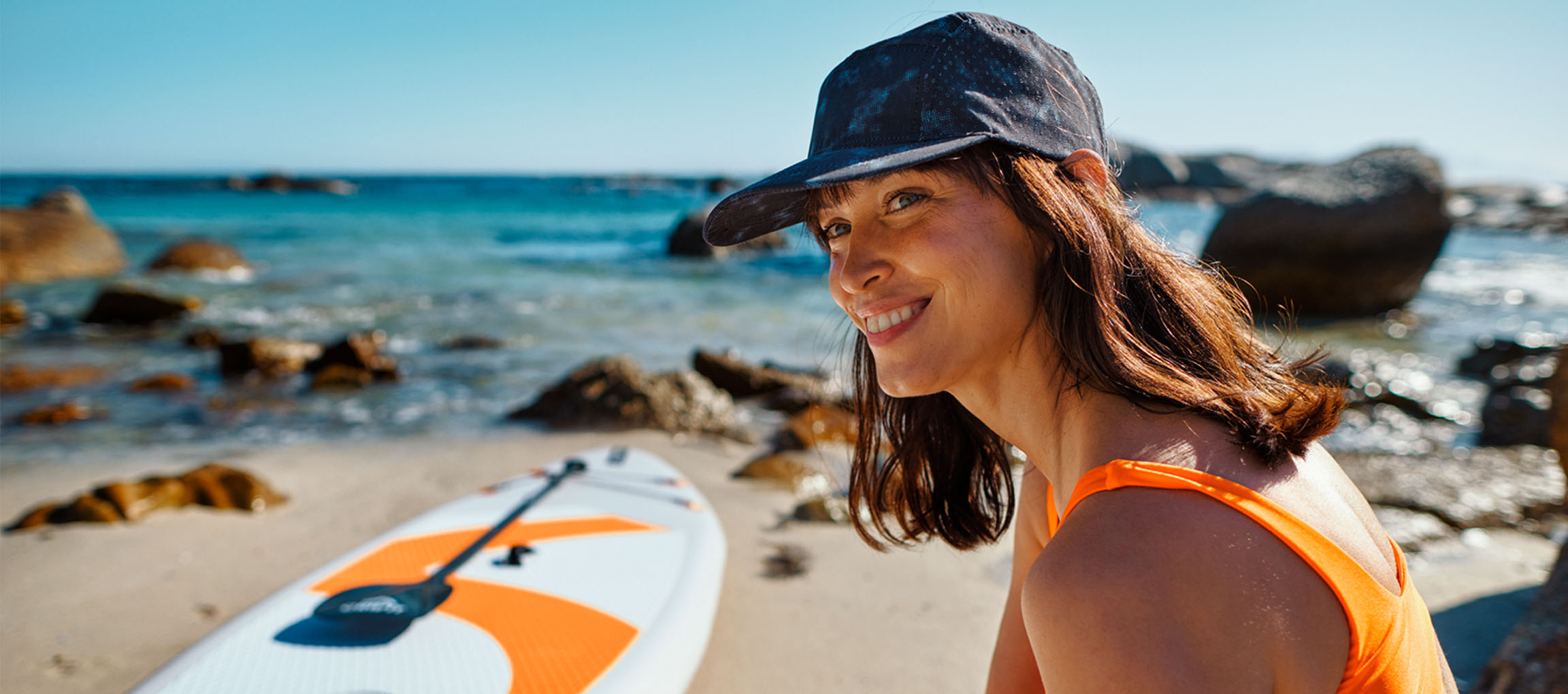 Smilende kvinde i orange badedragt og baseballkasket ved siden af et paddleboard på stranden.
