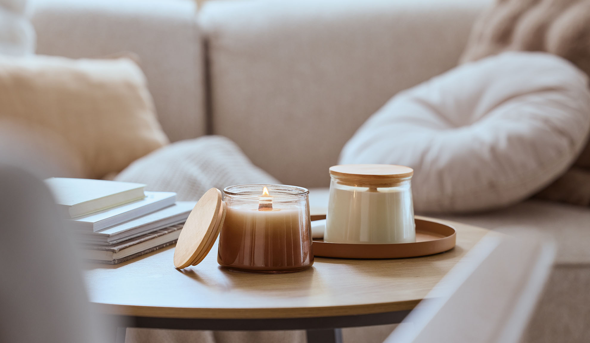 To duftlys på et træbord, det ene tændt, med en hyggelig sofa i baggrunden.