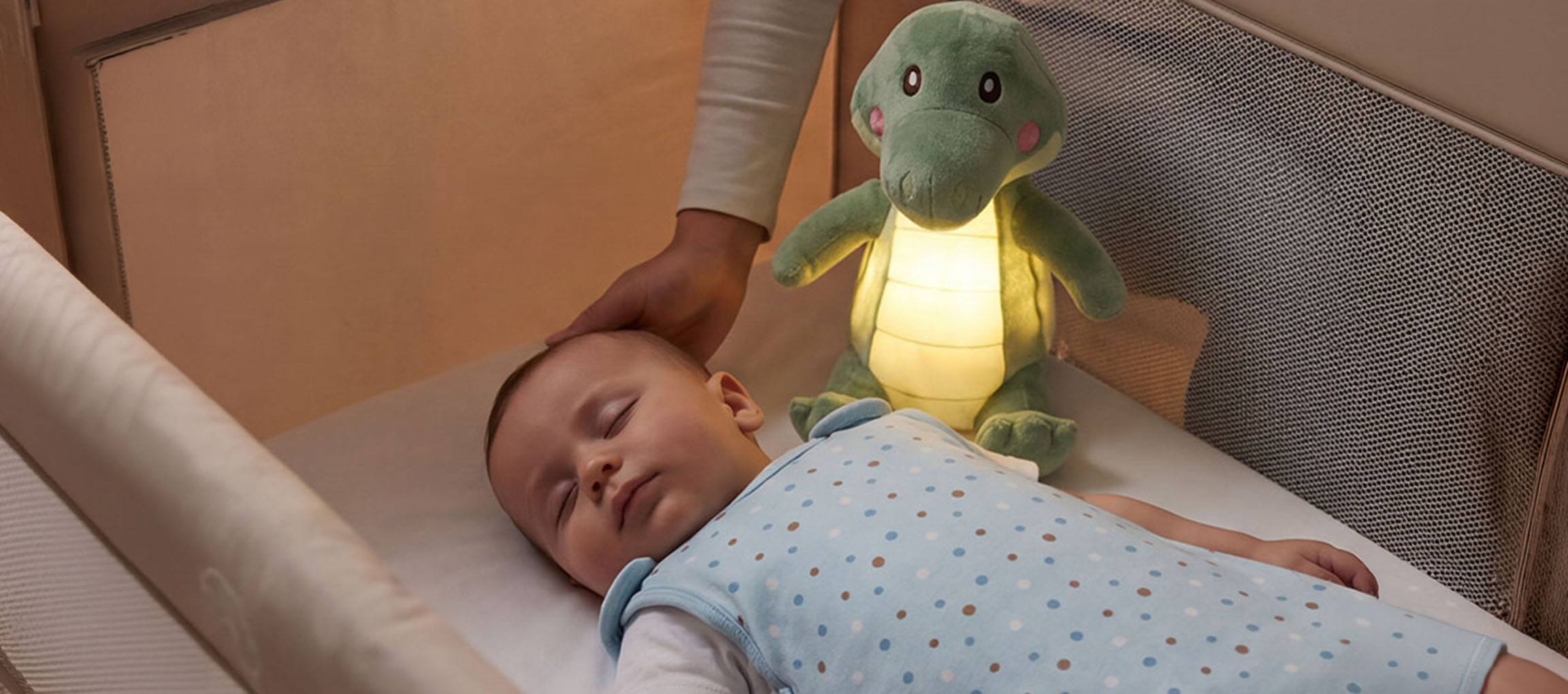Baby sover i tremmeseng med plys-natlampe, voksen aer babyens hoved.