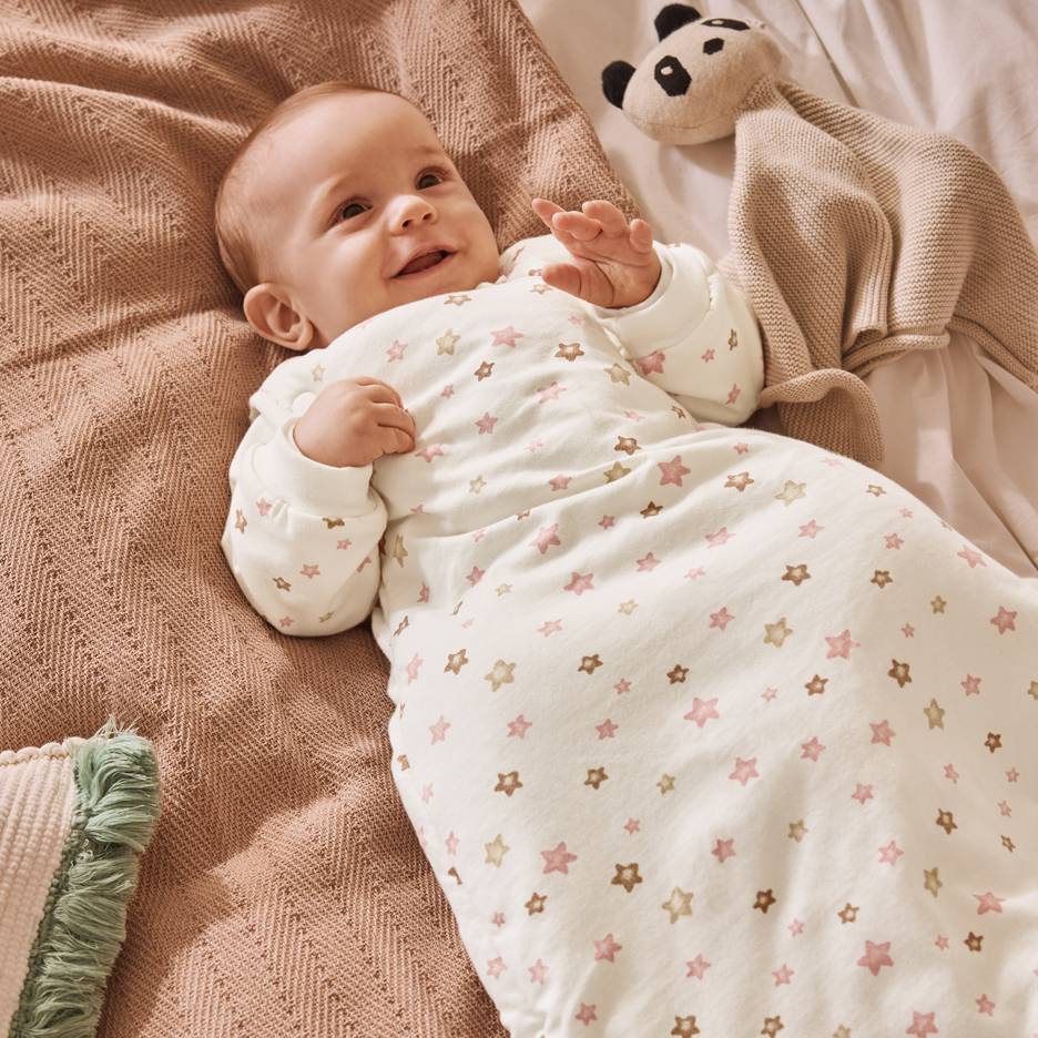 Smilende baby i hvid sovepose med lyserøde og brune stjerner, liggende på et tæppe.