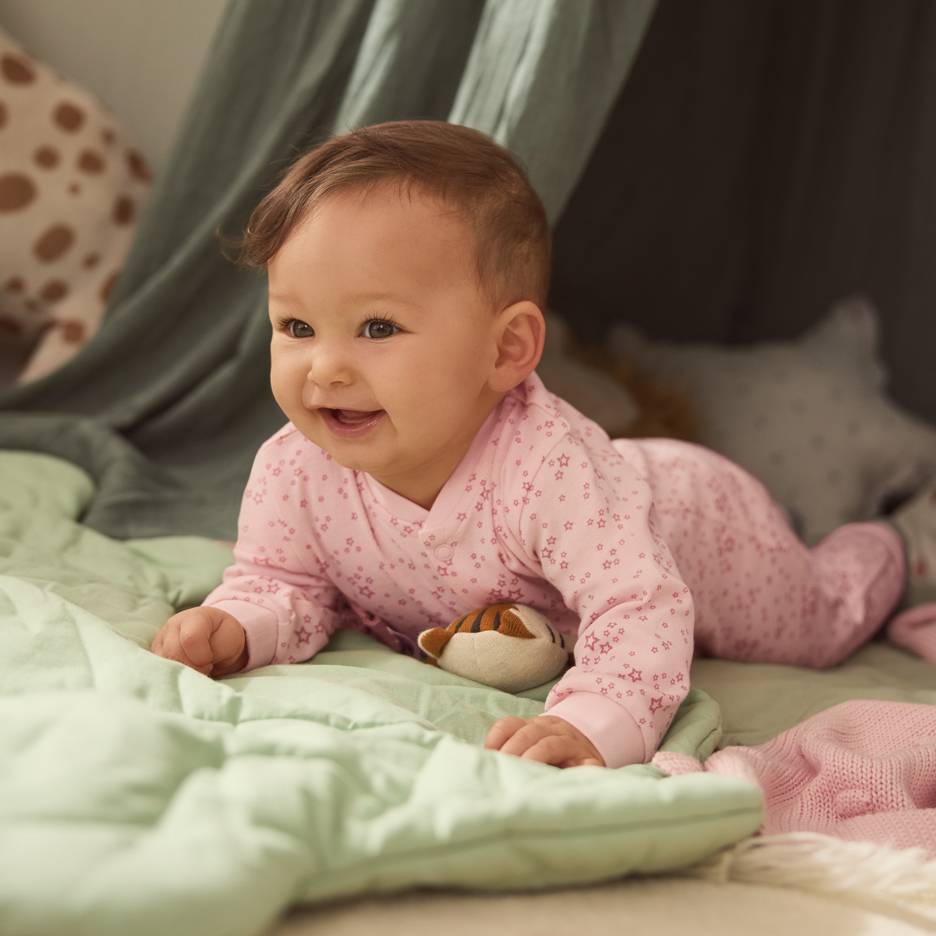 Baby i lyserød stjernepyjamas, liggende på et grønt tæppe med et plysdyr.